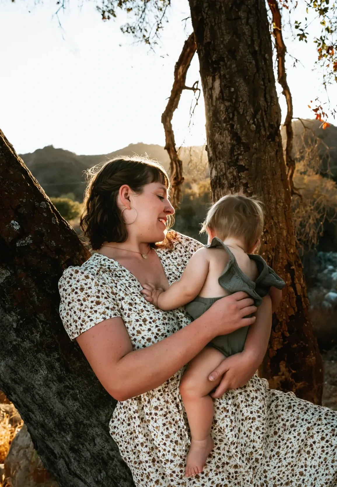 A woman sitting on a tree trunk holding a young child as they smile and enjoy an outdoor moment during sunset.