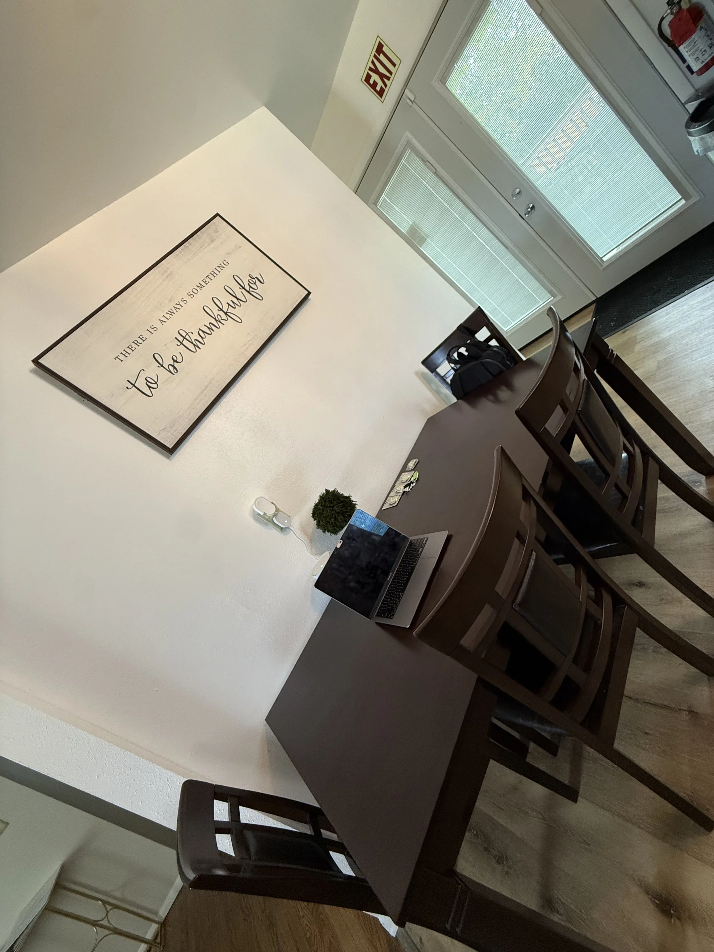 A dining area with a rectangular wooden table surrounded by six matching wooden chairs. A laptop and some small decorative items are on the table. On the wall, a framed sign reads 'There is always something to be thankful for'. There are glass doors 