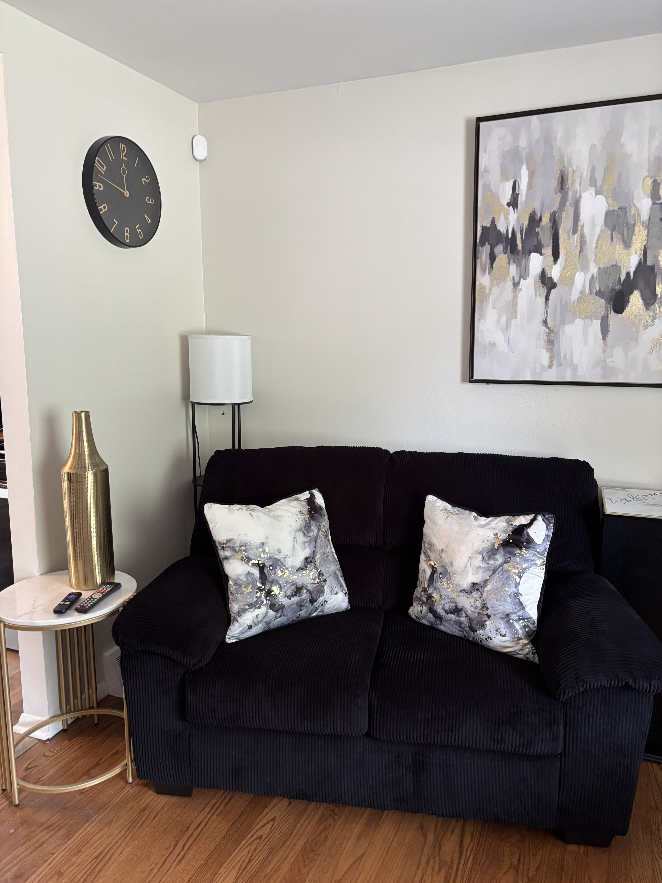 A living room corner with a black sofa, decorative pillows, a side table with two remote controls and a gold vase, a white floor lamp, a wall clock, and a large abstract painting on the wall.