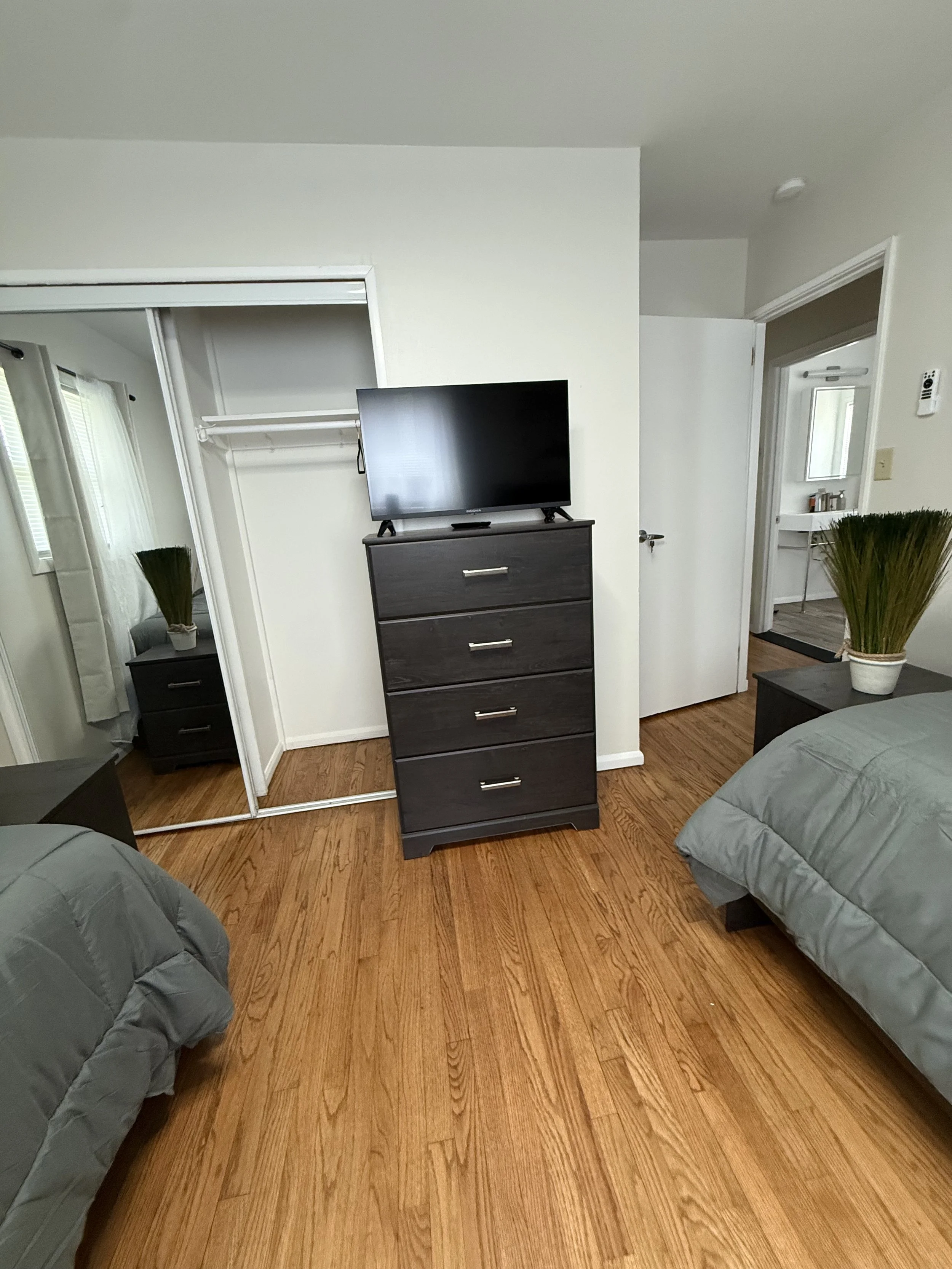A bedroom with wooden flooring, two beds with gray bedding, a black dresser with a TV on top, a mirrored closet door, a plant in a white pot, and an open doorway leading to a bathroom with a mirror and counter.