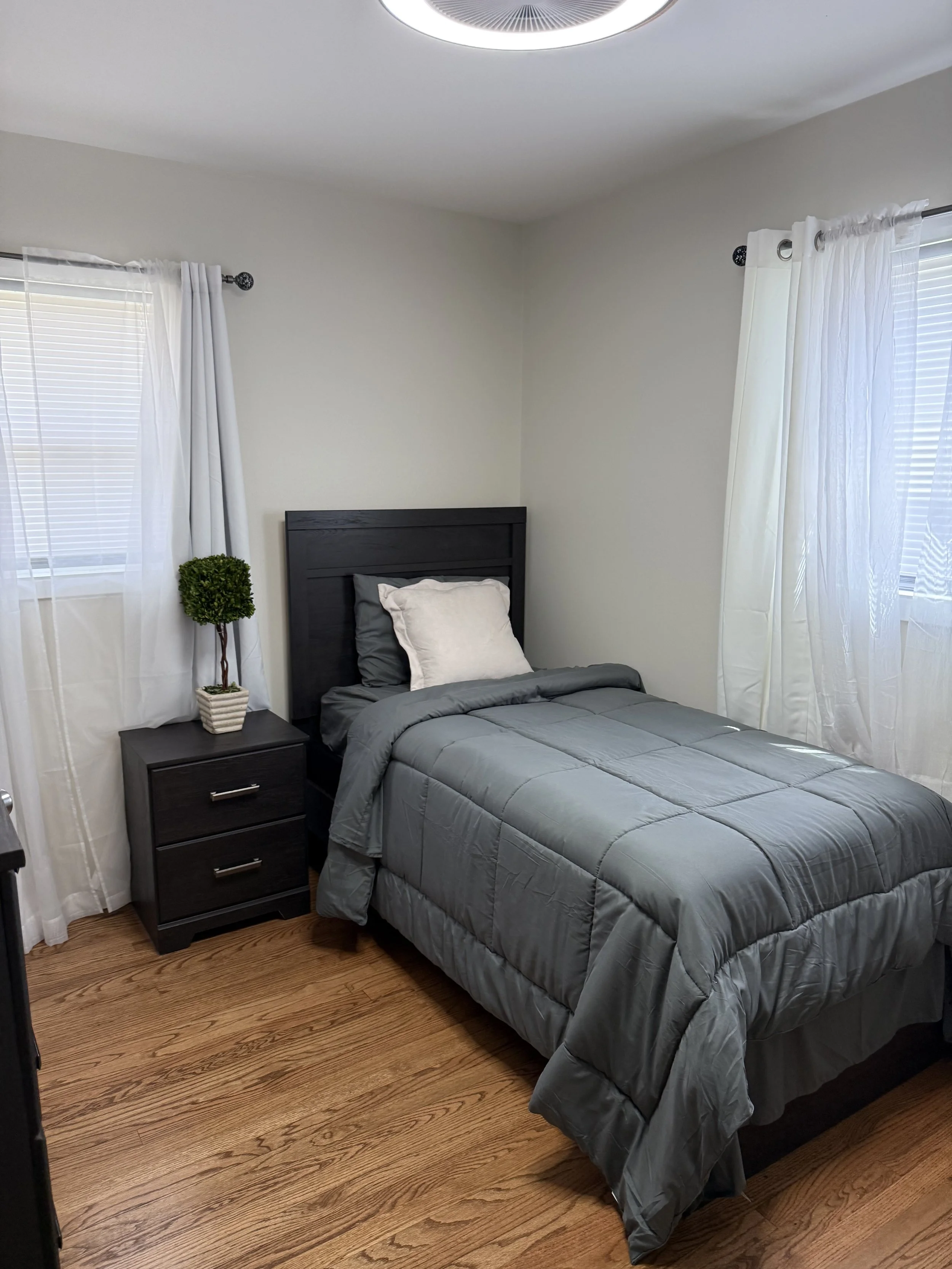 A bedroom with a bed, nightstand, and two windows with white curtains. The bed has a dark headboard, gray bedding, and two pillows. There is a small potted plant on the nightstand. The room has wooden flooring and a ceiling light fixture.