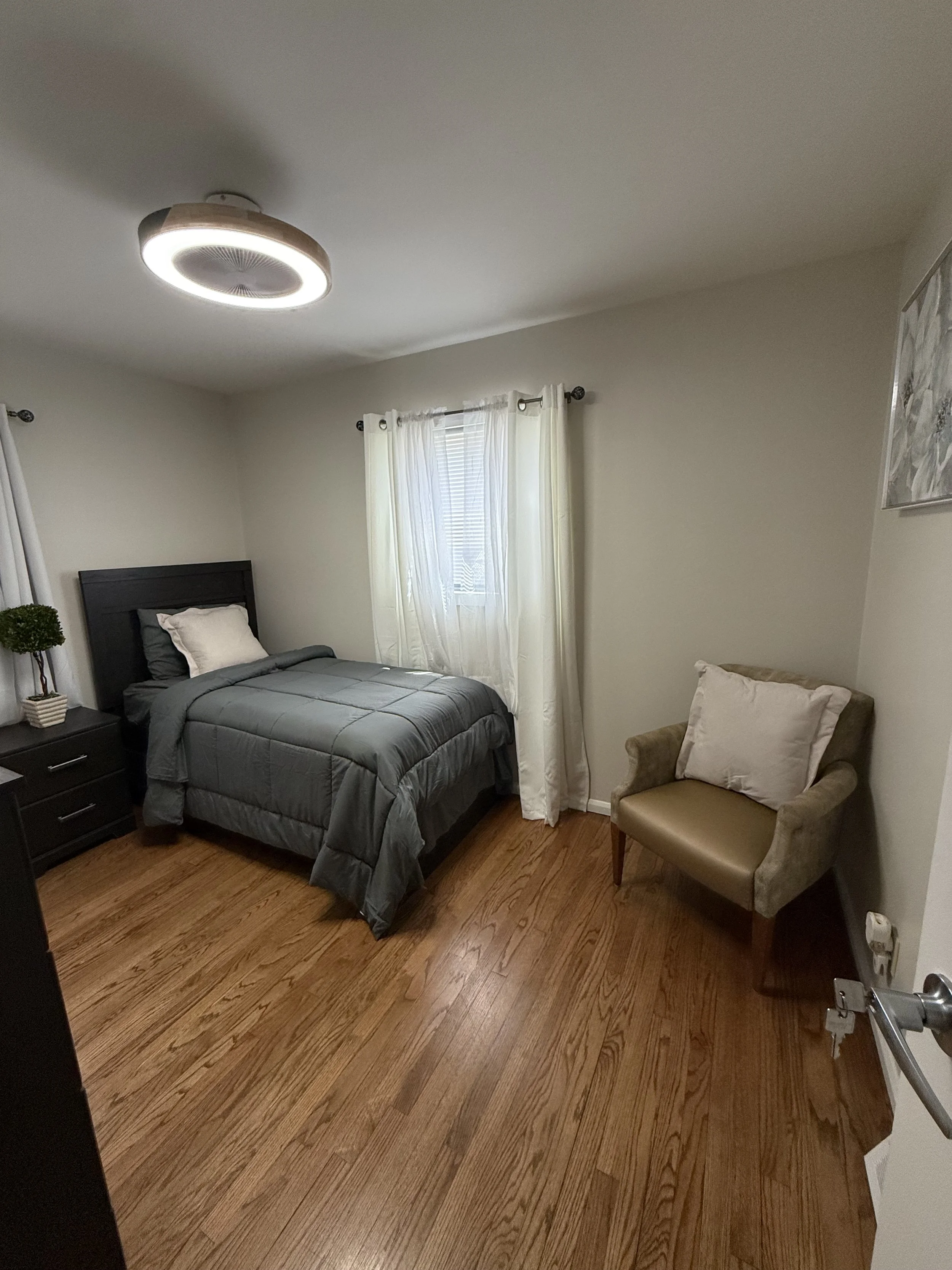 A bedroom with a bed covered in a gray quilt, a black headboard, white pillows, a small black nightstand with a potted plant, a chair with a pillow, a window with white curtains, and hardwood floors.