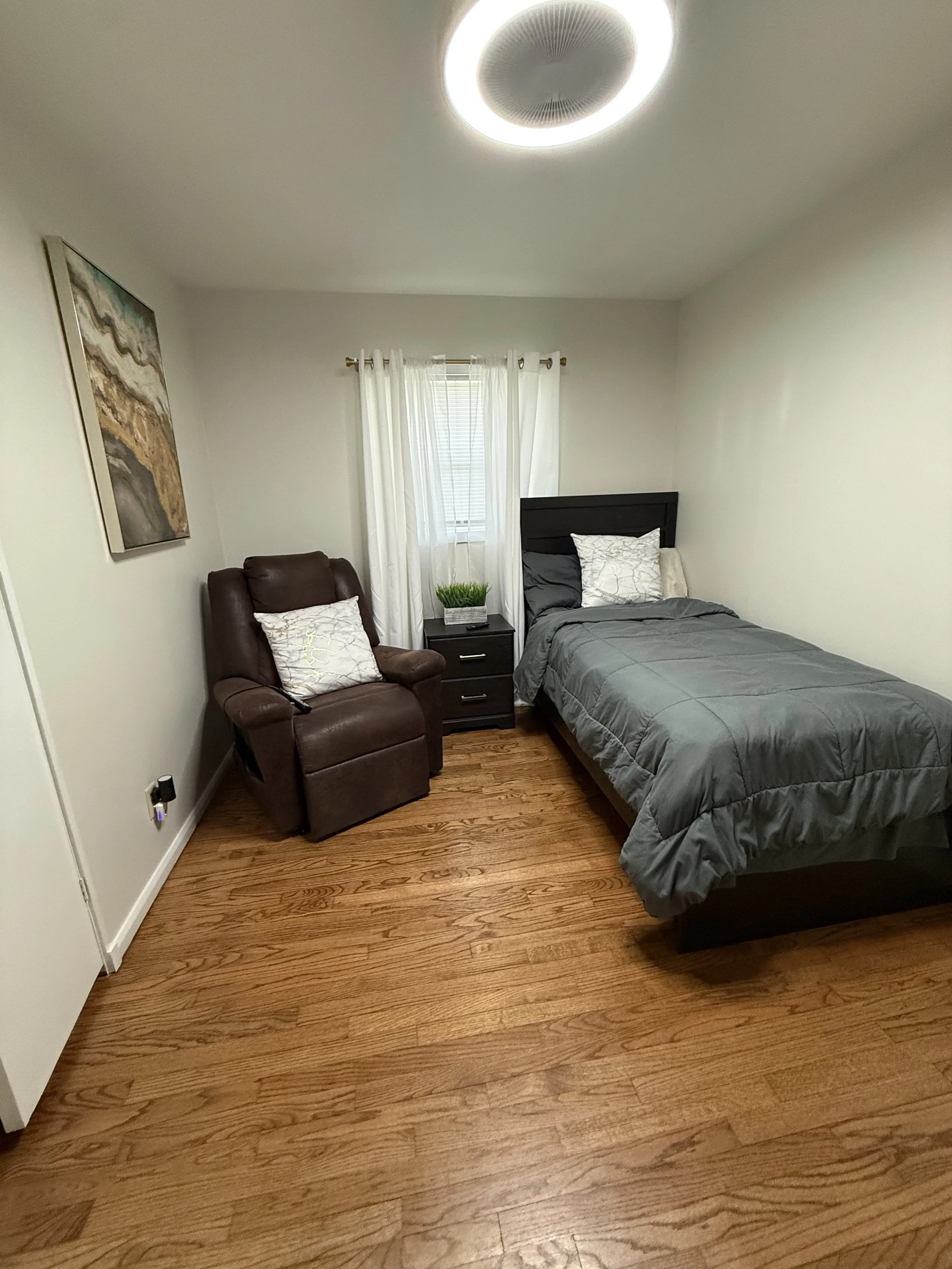 A bedroom with hardwood floors, a bed with gray bedding and pillows, a dark wooden nightstand, a brown leather armchair with a decorative pillow, a window with white curtains, a wall art piece, and a ceiling light fixture.