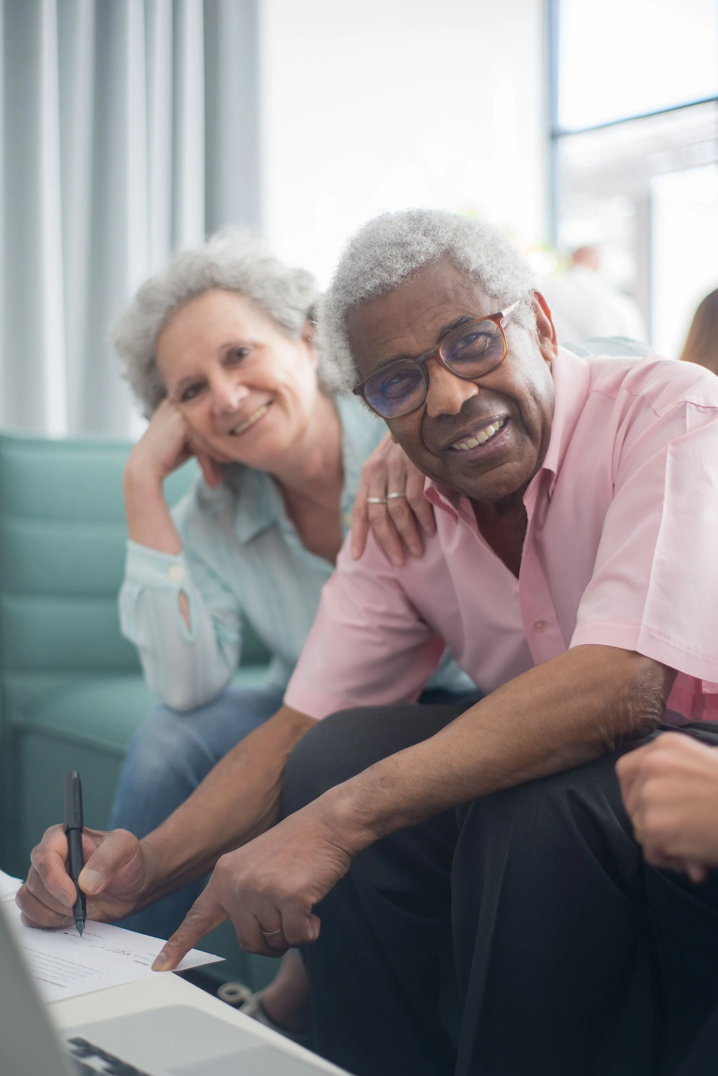 An elderly man with glasses leaning on a table, pointing to a paper with his finger, while a smiling elderly woman watches. They are in a bright room, possibly engaging in a social or educational activity.