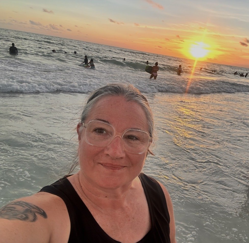 A woman wearing glasses and a black tank top takes a selfie at the beach during sunset, with people swimming and playing in the water in the background.