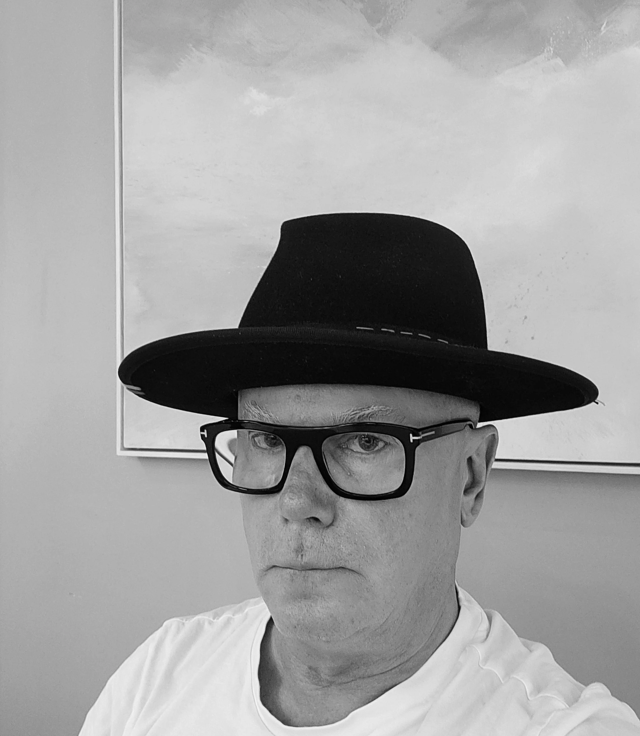 A black and white photograph of a person wearing glasses and a hat, with the person’s face and upper body visible. The individual is turned slightly to the right, with a neutral expression, and is wearing a light-colored shirt. The background is plain, with a framed picture or window behind them.