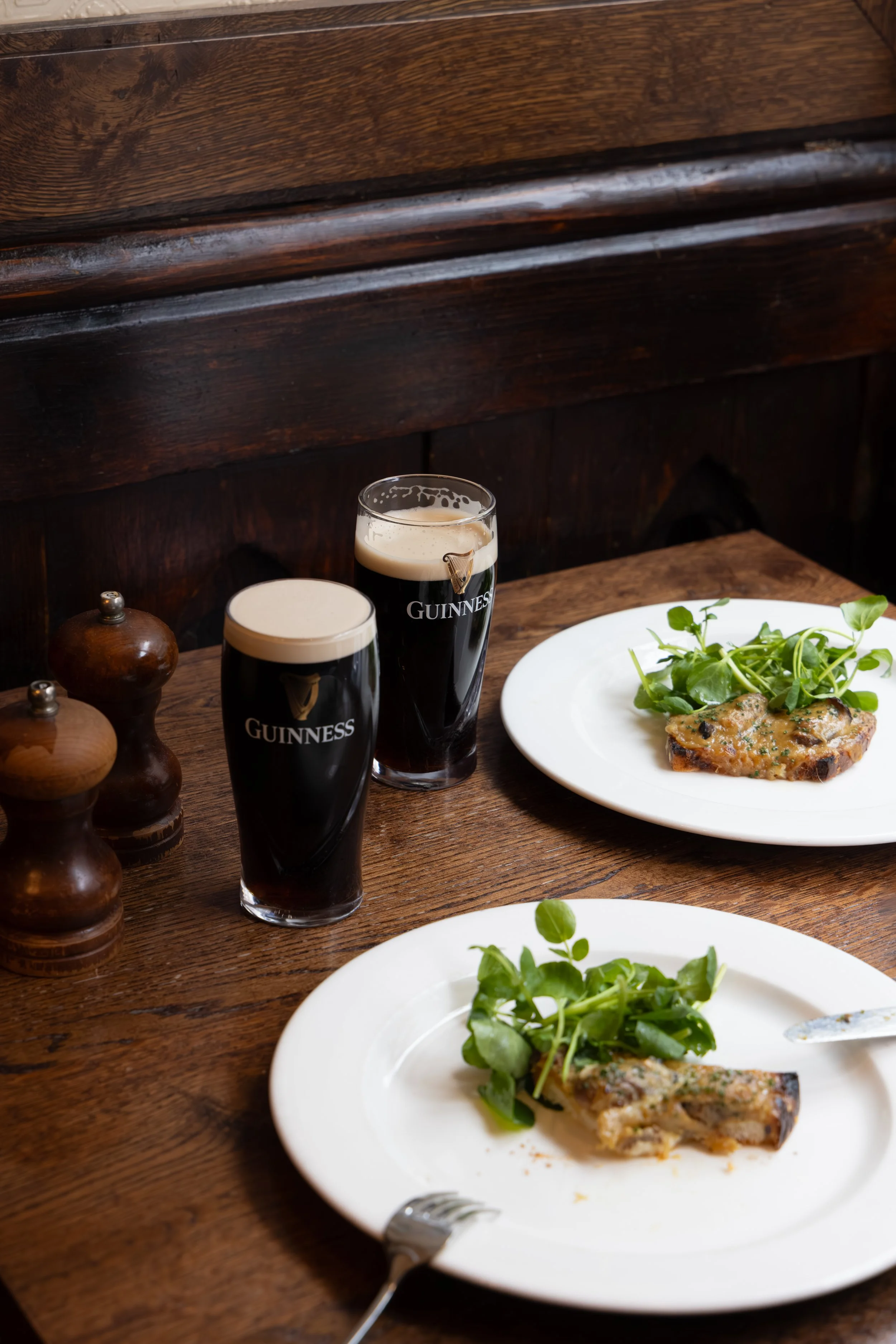 Two pints of Guinness beer and a white plate with grilled fish topped with green herbs and a side of fresh salad on a wooden table.