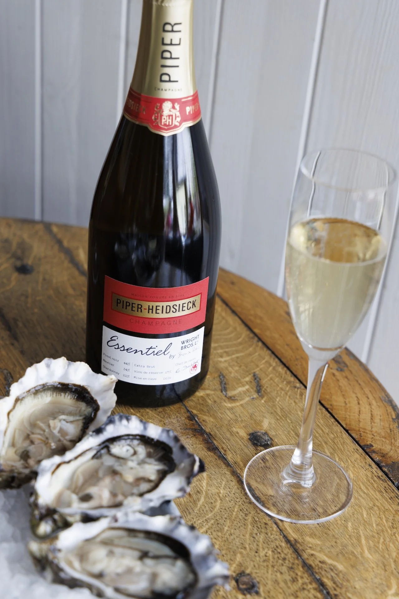 A bottle of Piper-Heidsieck champagne, a filled champagne flute, and oysters on a wooden table with a white paneled wall in the background.