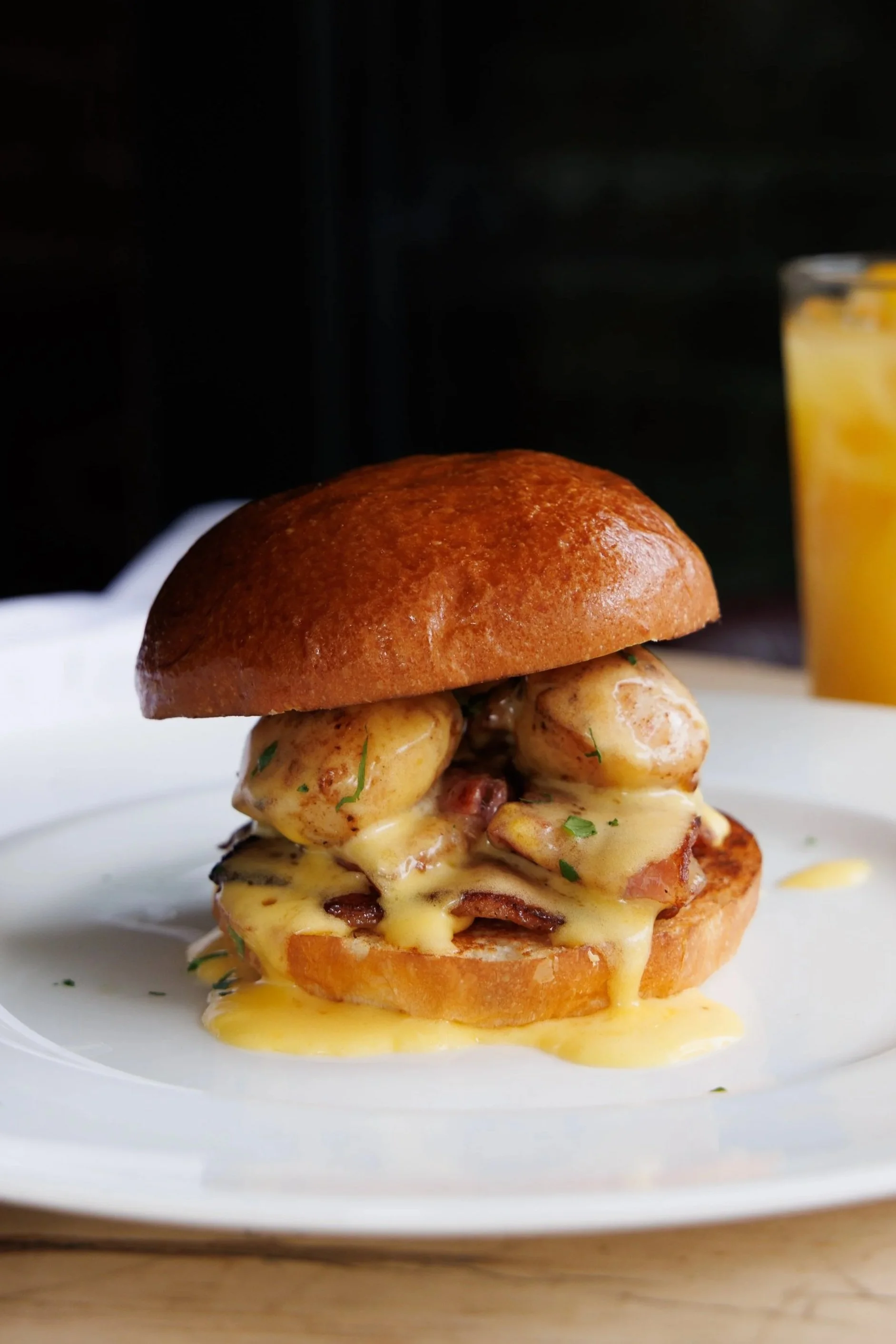 Close-up of a gourmet sandwich with melted cheese, grilled chicken, and herbs on a white plate. In the background, a glass of orange juice.