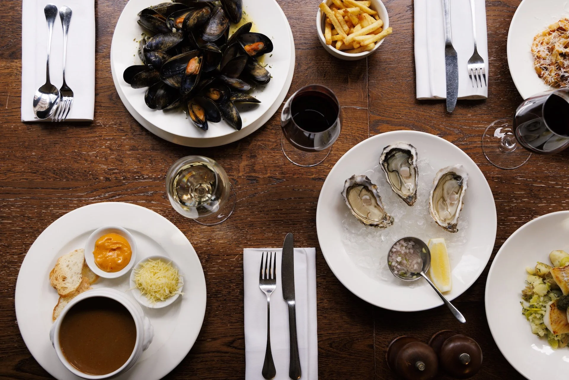 A top-down view of a dinner table set with various seafood dishes, including several oysters on the half shell, steamed mussels, French fries, a bread roll with cheese and a soft-boiled egg, a plate of pasta with grated cheese, and glasses of red win