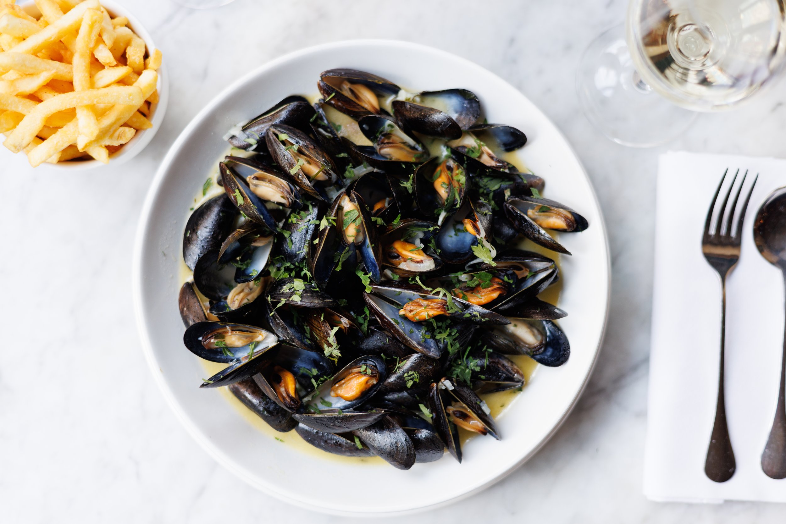 A white plate with cooked mussels garnished with chopped parsley, served with a side of French fries and a glass of white wine.