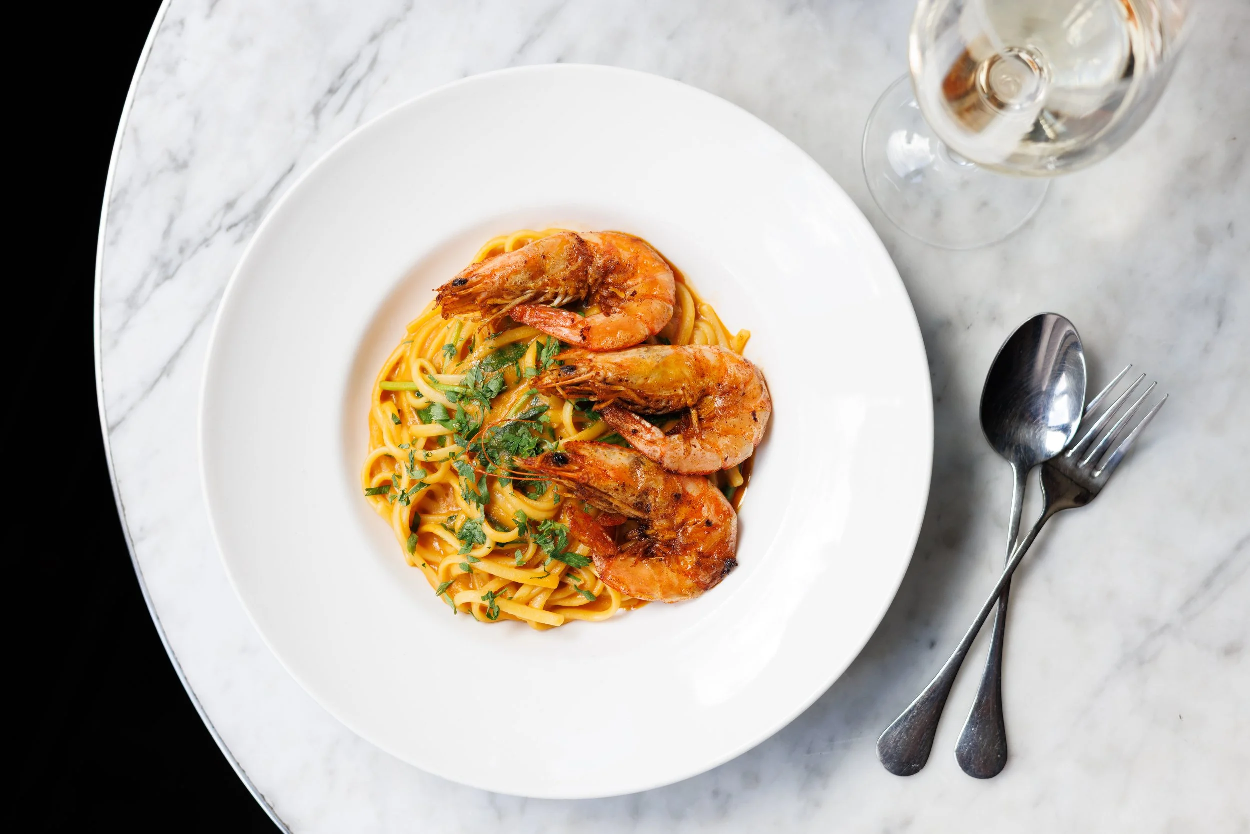 Plate of spaghetti with shrimp and herbs, served with a glass of white wine on a marble table.