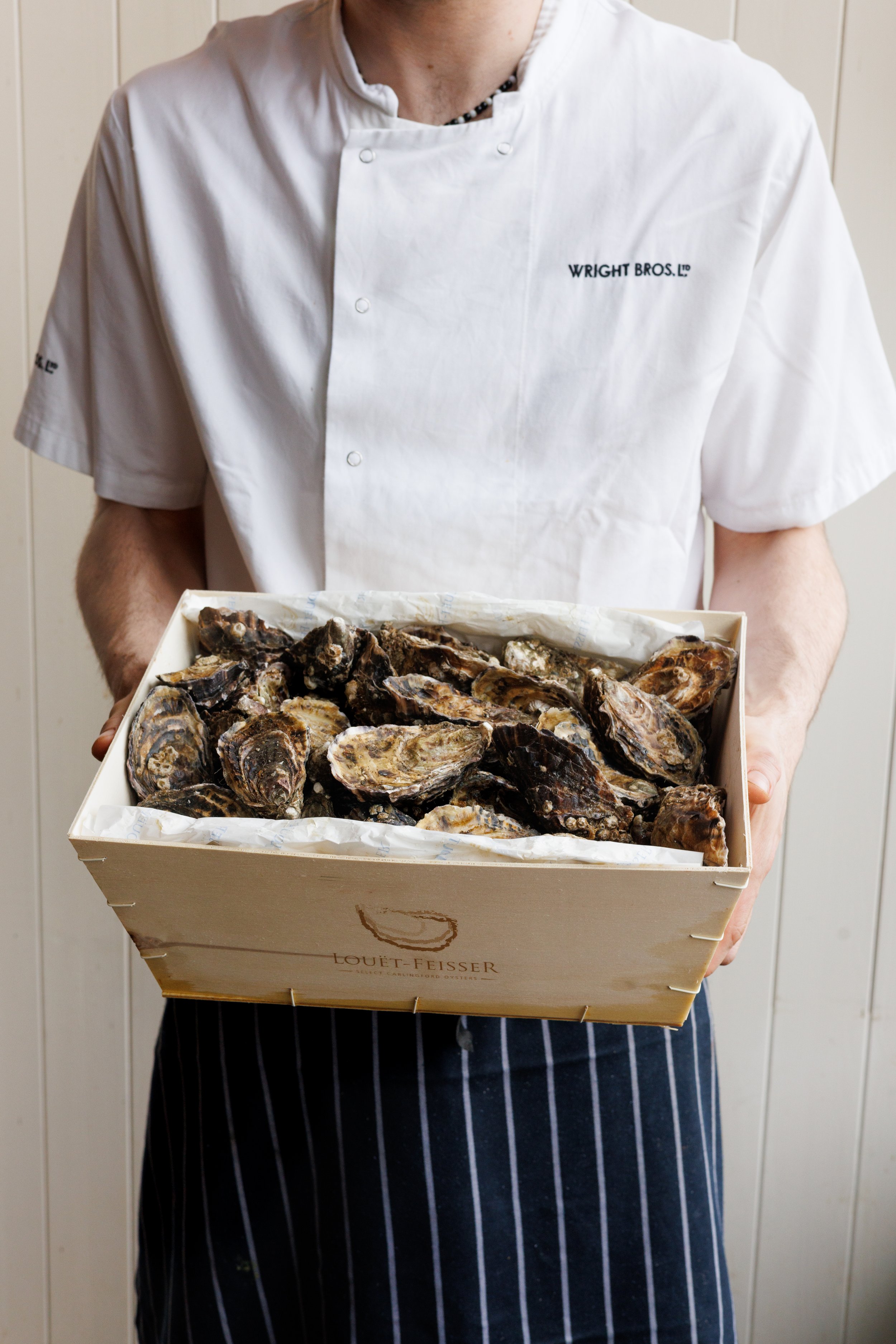 A person wearing a white apron with 'Wright Bros.' embroidered on it, holding a crate of fresh oysters. The person is standing against a plain, light-colored wall.