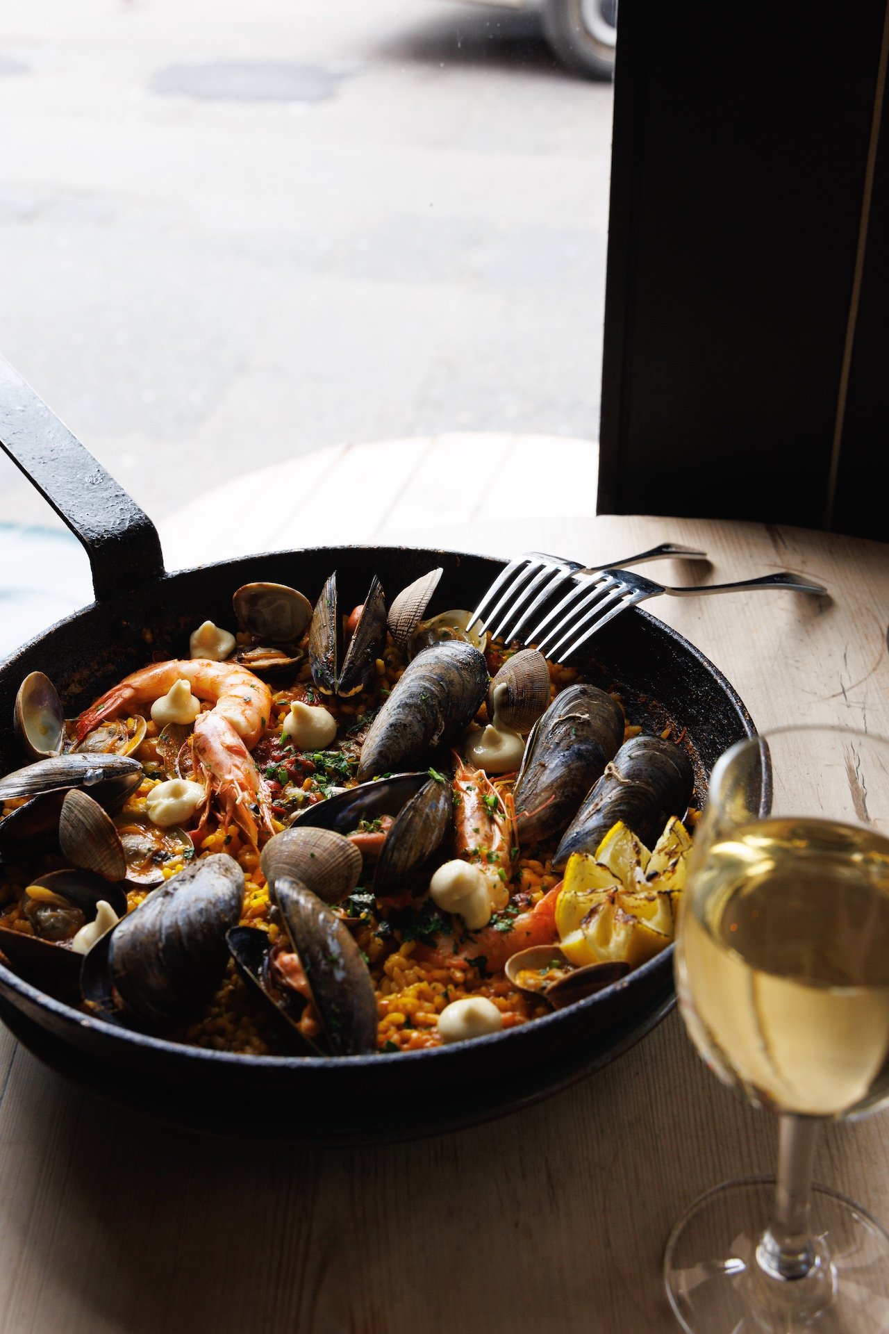 Seafood paella in a black skillet with shrimp, mussels, clams, and vegetables, served with a glass of white wine on a wooden table near a window.