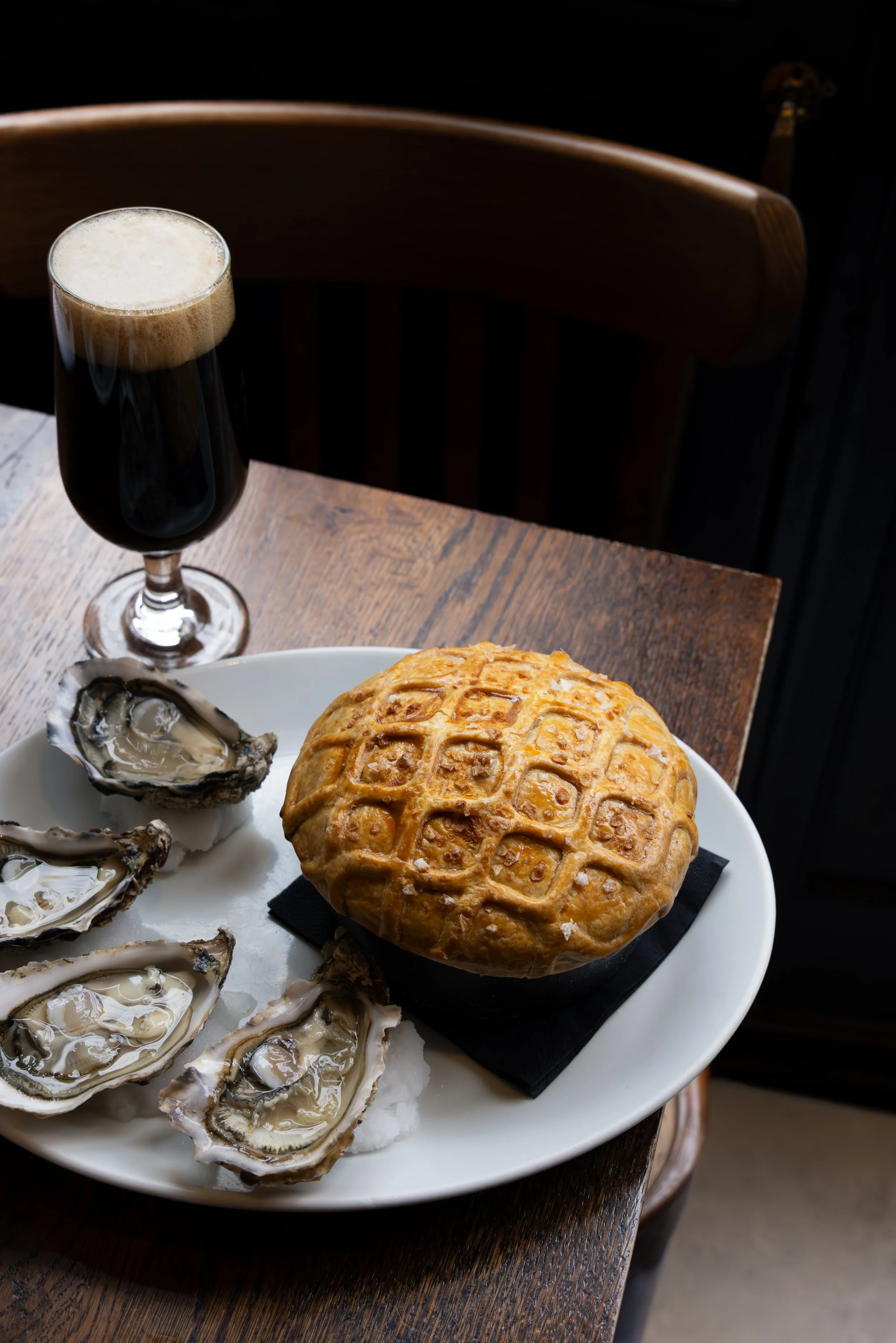 A white plate of fresh oysters on ice, a round baked dish with a lattice crust, and a tall glass of dark stout beer with foam, on a wooden table.