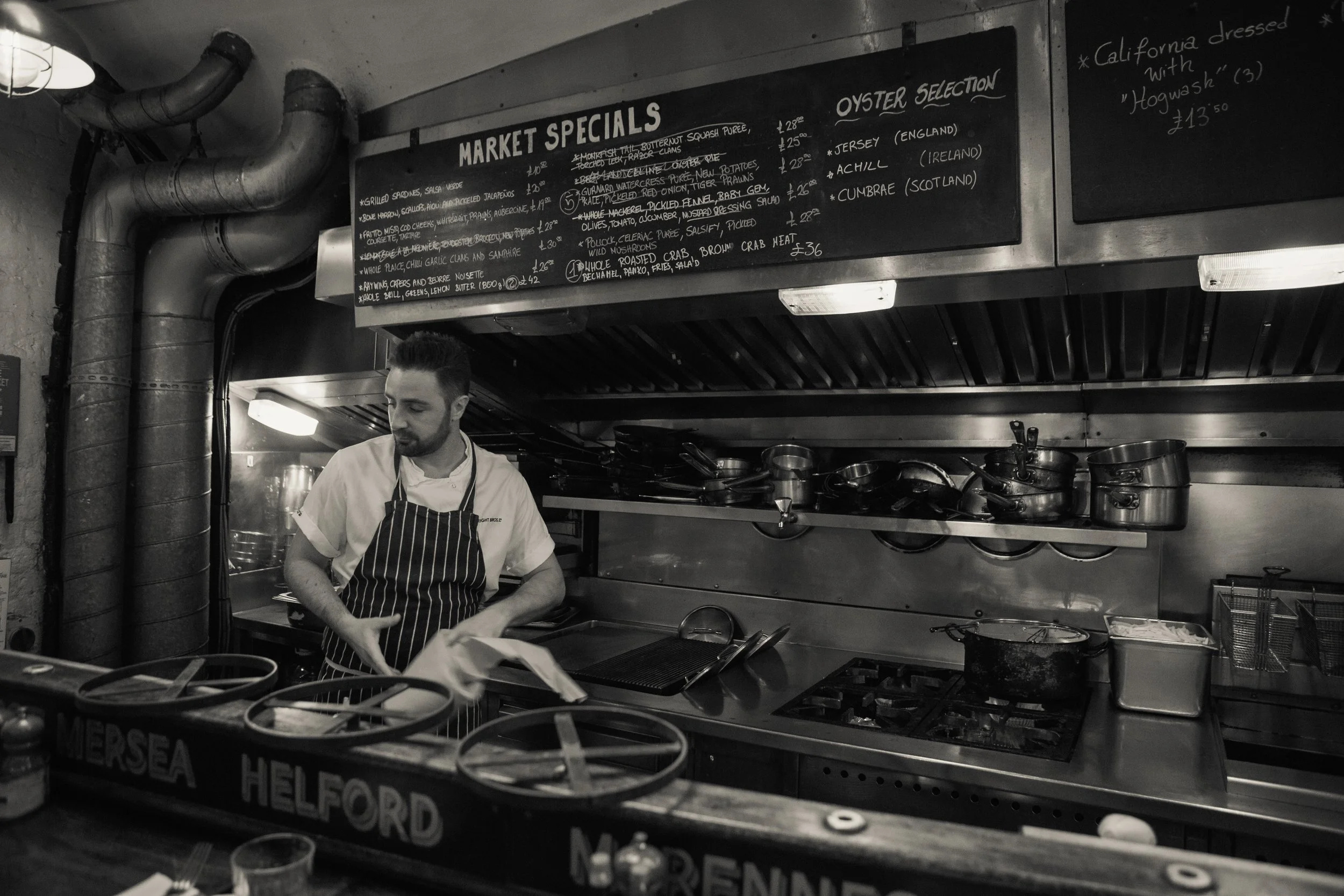 A man in a striped apron preparing food behind a counter at a restaurant or market, with a chalkboard menu listing market specials and oyster selections above him.