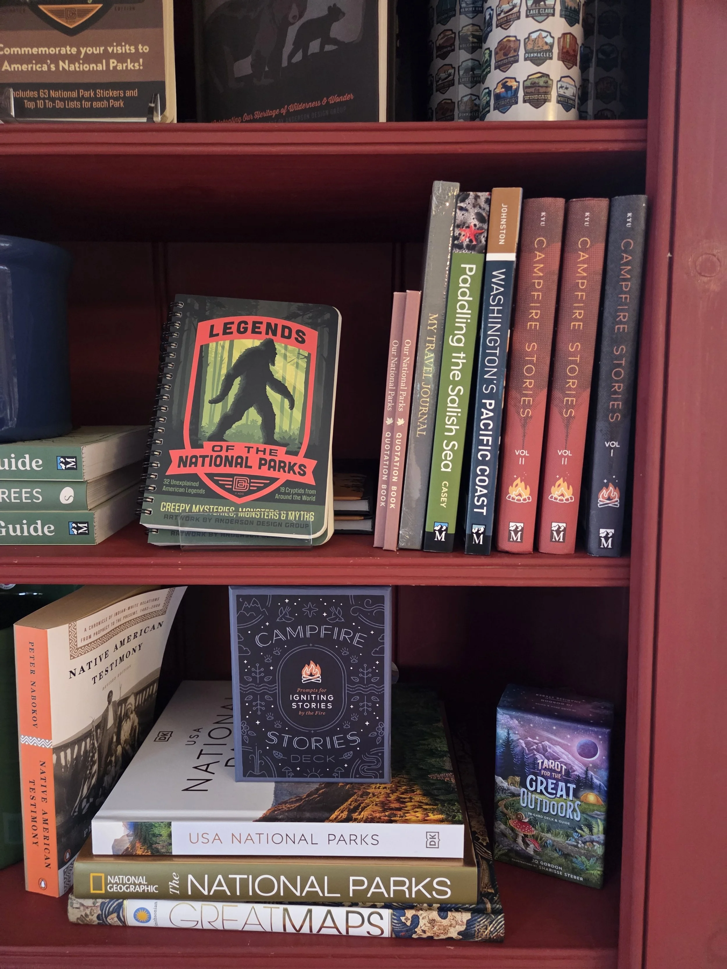 Bookshelf containing books about national parks, wilderness stories, camping, and outdoor adventures. Notable titles include 'Legends of the National Parks,' 'Walshing the Seas,' 'Paddling the Solish Sea,' 'My Travel Journal,' 'Our National Parks,' 'Campfire Stories,' 'Great Maps,' and 'Tarot for the Great Outdoors.'