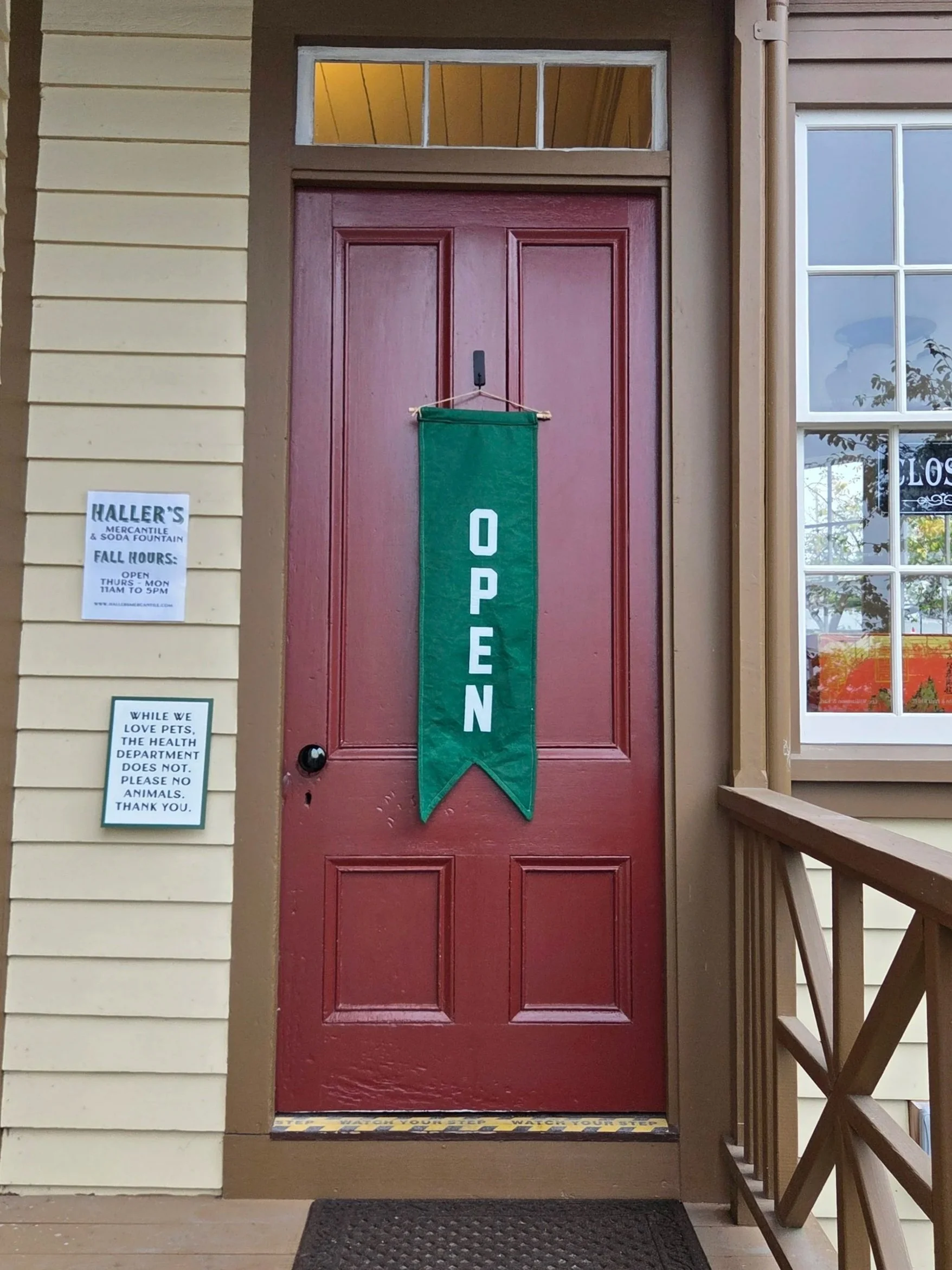 Red front door with a large green 'OPEN' banner hanging in the center, flanked by signs on either side, on a porch with wooden flooring and stairs, part of a yellow house with beige trim.