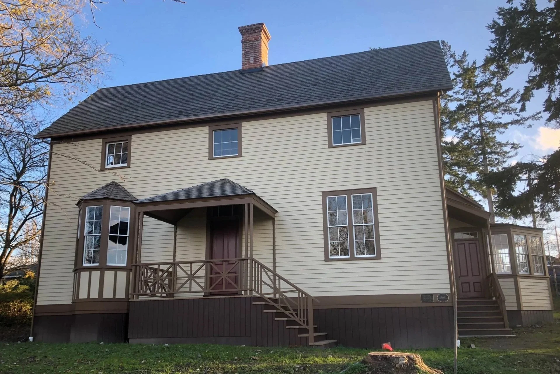 A two-story house with beige siding, brown trim, and a dark grey roof. It has multiple windows and an elevated porch with stairs leading to the front door. Surrounding trees and a clear blue sky are in the background.