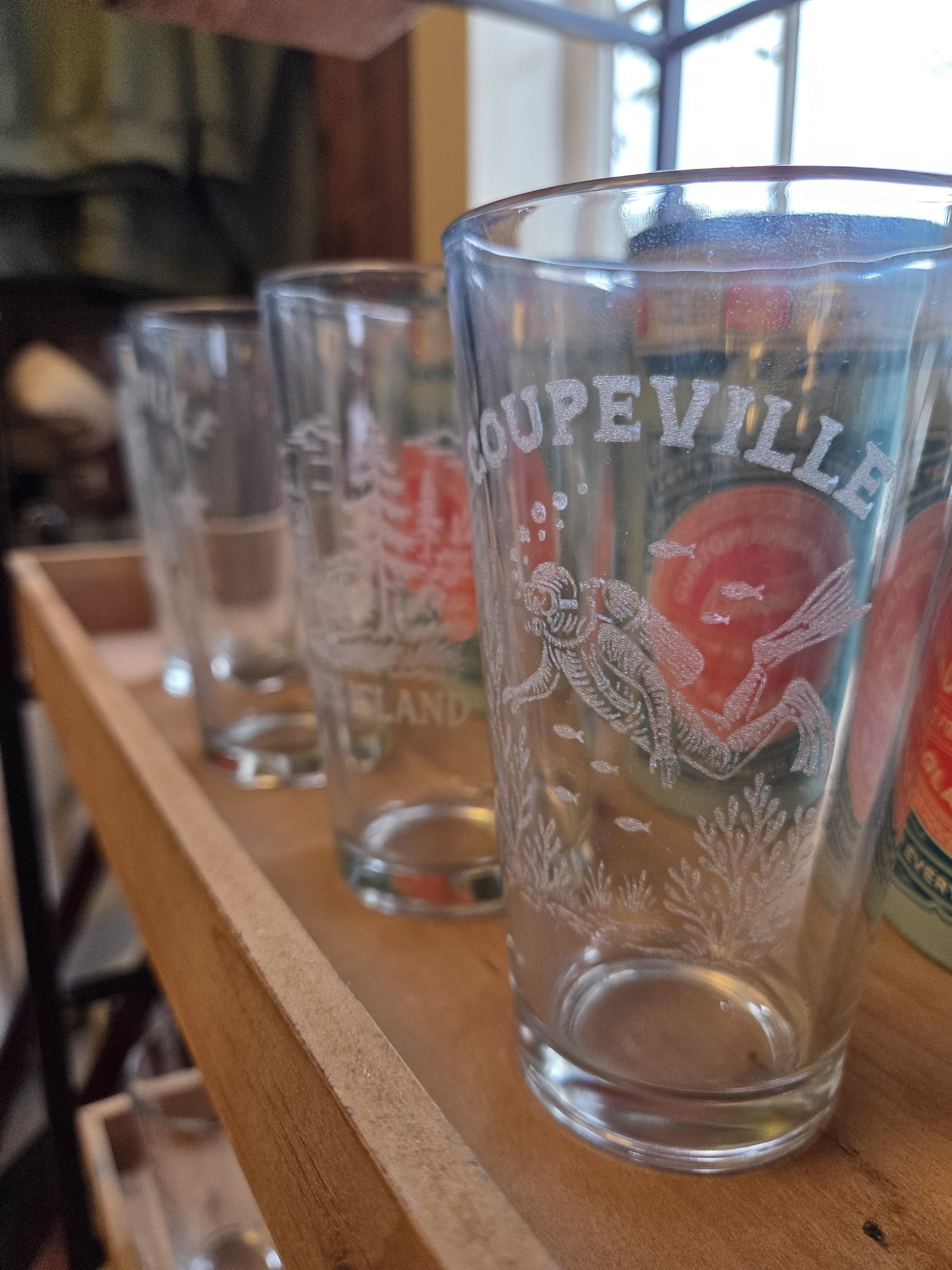 Three clear drinking glasses on a wooden shelf, with the front glass featuring a design of a scuba diver and the word 'COUPEVILLE'.