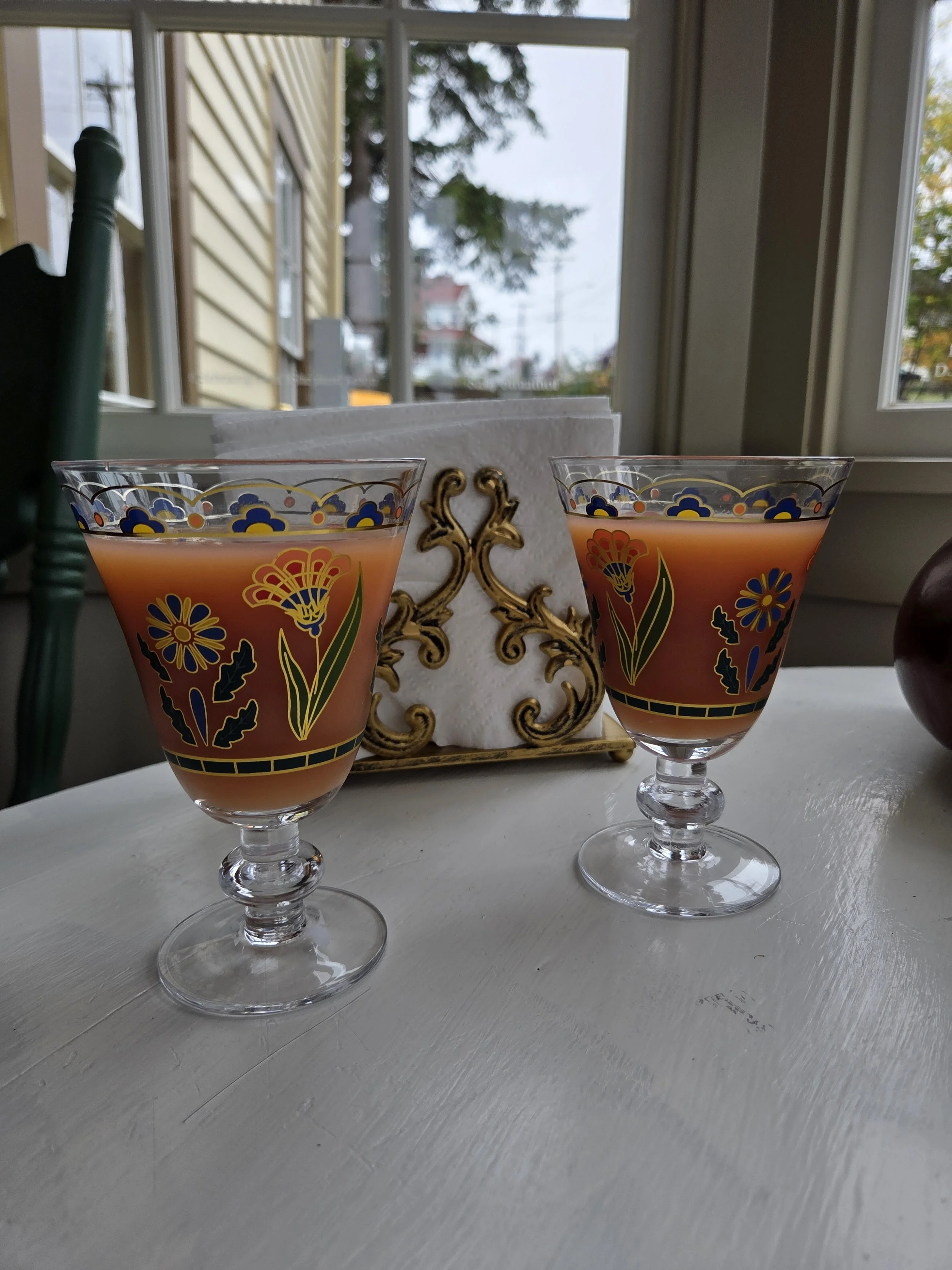 A pink drink in glass goblets decorated with floral and blue patterns sit on a white table, with a decorative napkin holder between them. A window showing trees and houses is in the background.