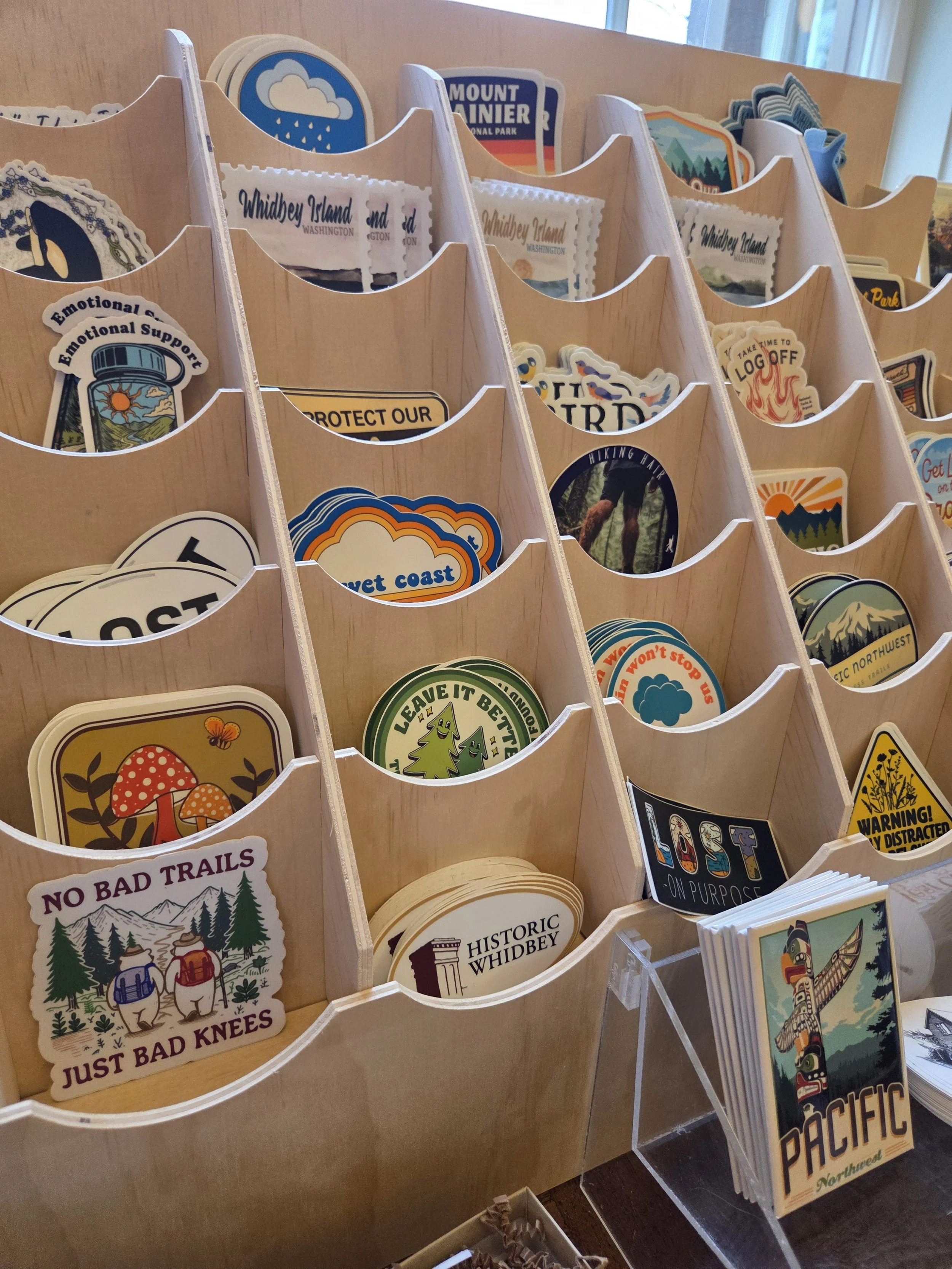 Wooden rack filled with various colorful stickers and patches depicting nature, travel, and outdoor themes, such as mountains, forests, and animals, displayed on a tabletop near a window.