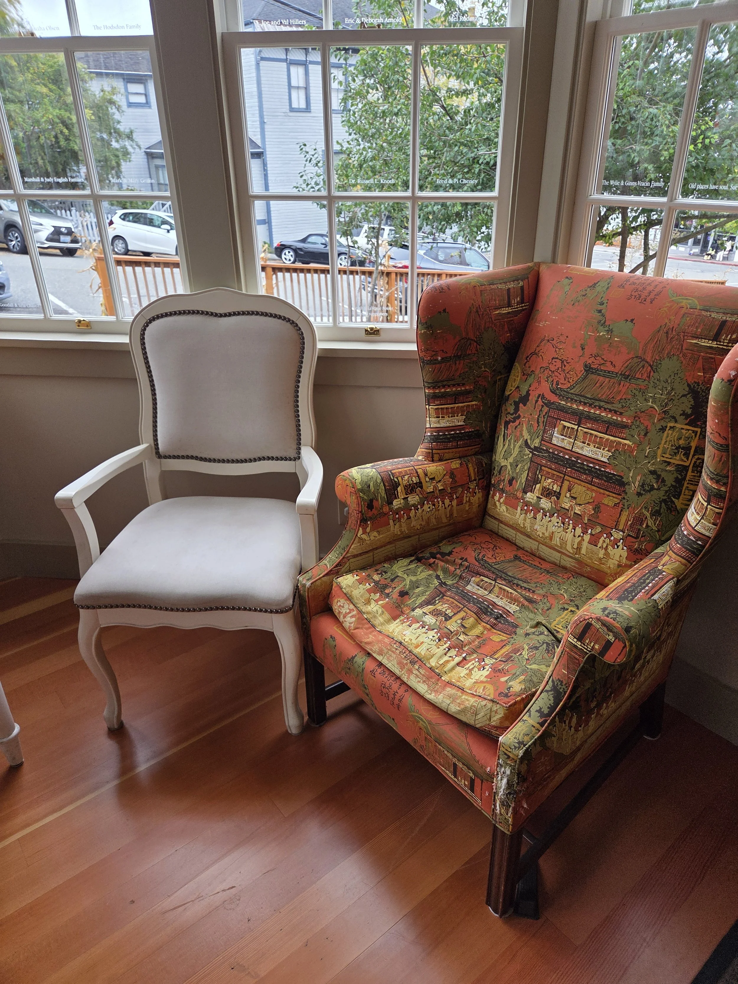 A white chair and a colorful patterned armchair are positioned near a large window with a view of car-lined small-town street surrounded by trees.