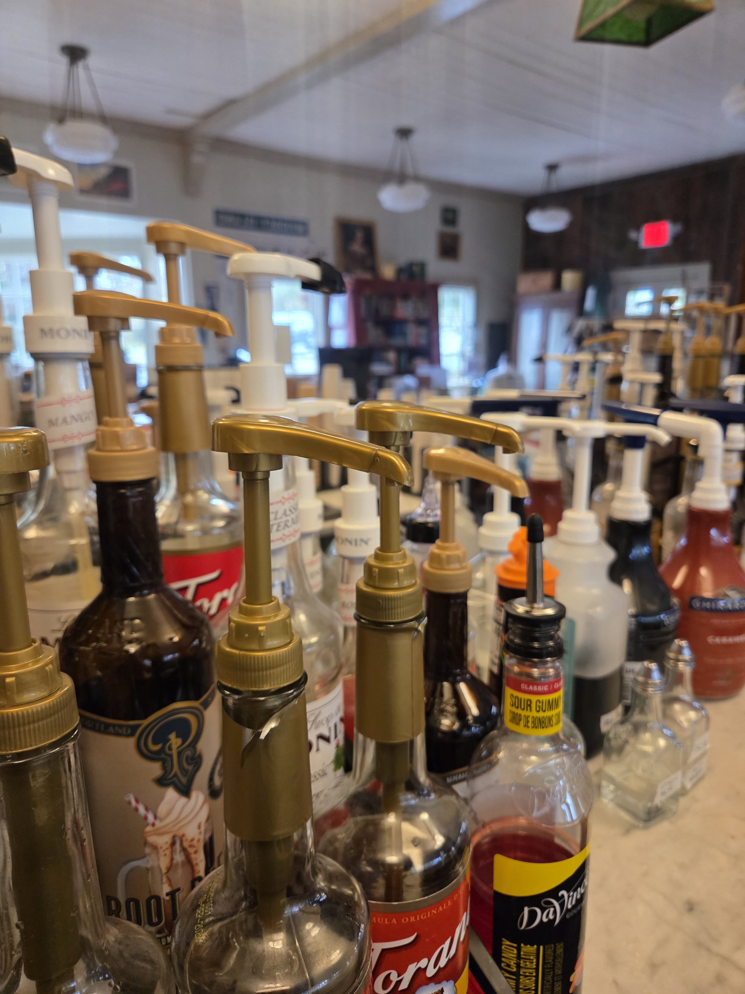 Multiple bottles of flavored syrup with pump dispensers on a table, with a blurred background of a room with hanging lights, windows, and shelves.