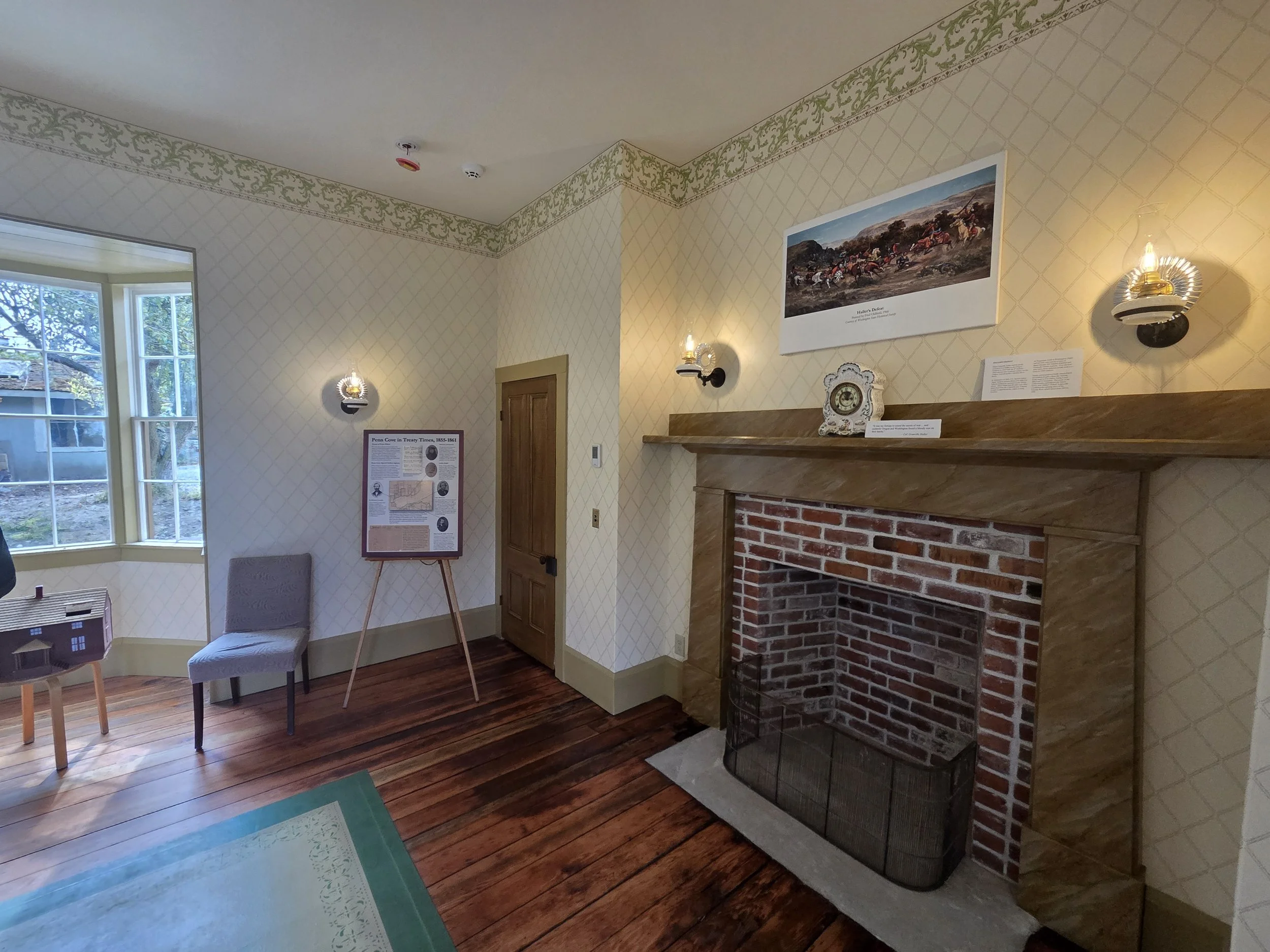 The image shows the interior of a room with a fireplace, wall-mounted lamps, a wooden door, and framed artwork on the walls. There is a large window with a view of trees outside, and the room has wood flooring and patterned wallpaper.