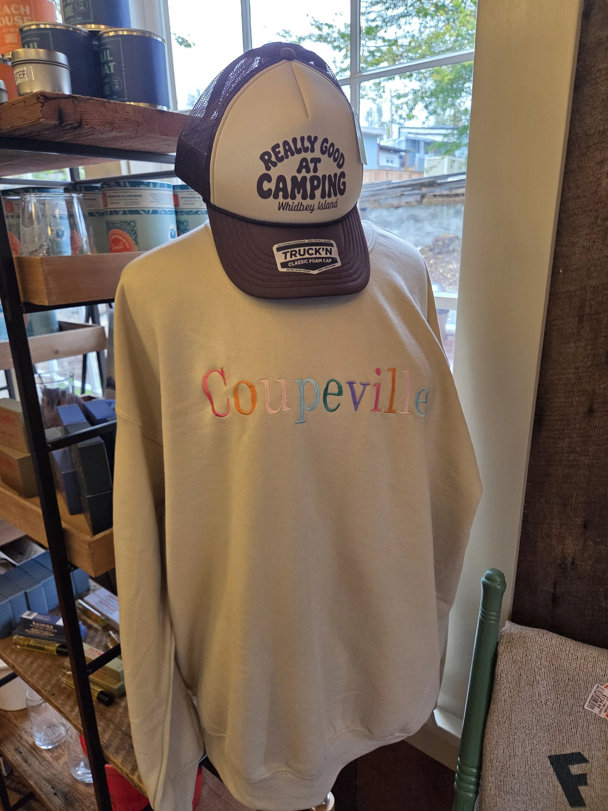 Cream-colored sweatshirt with multicolored embroidered text reading 'Coupville' displayed on a mannequin. A trucker hat with a mesh back, featuring a label that says 'Really good at camping Whidbey Island,' is placed on top of the sweatshirt. The background shows shelves with can near a window with trees outside.