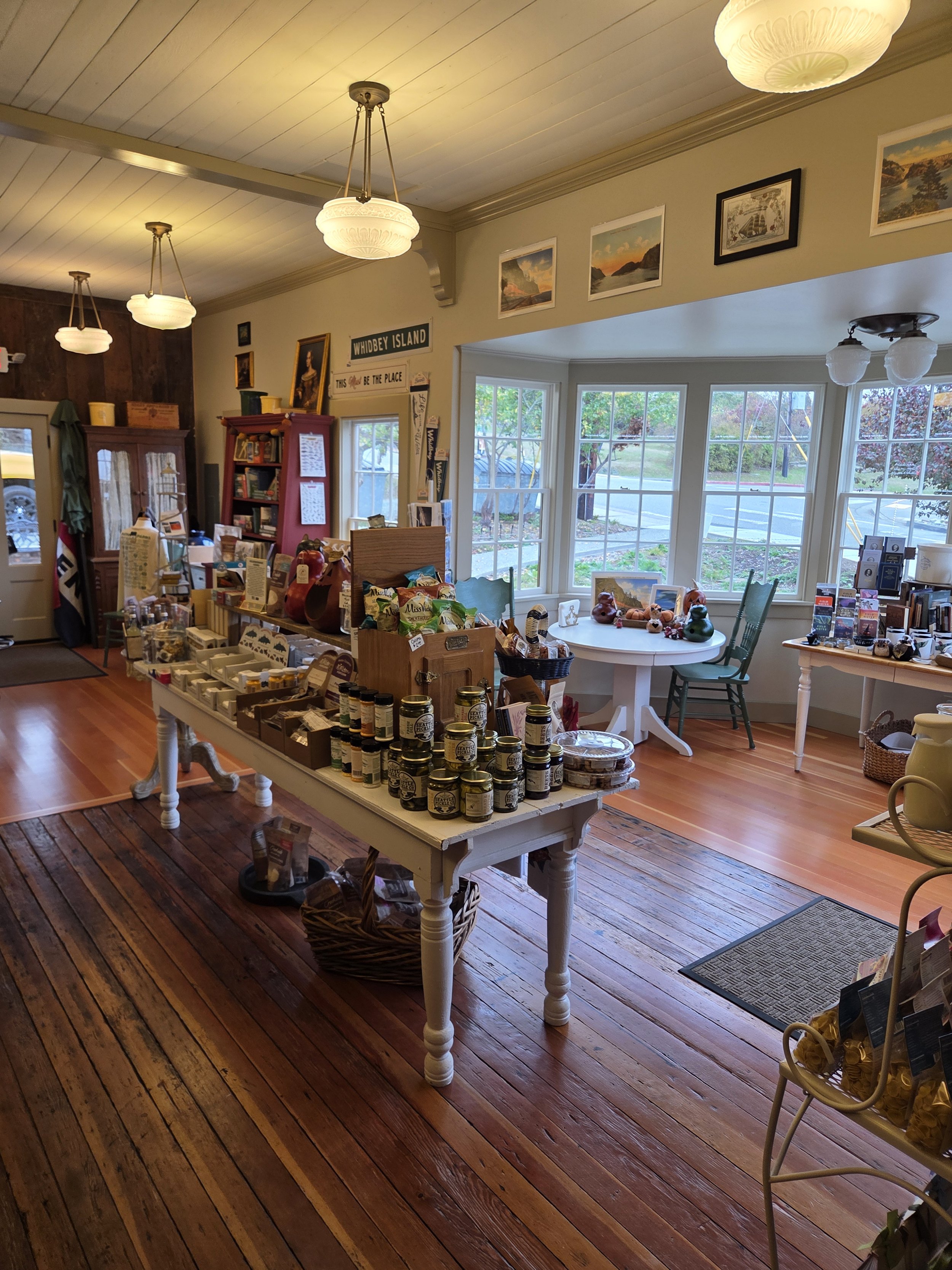 Interior of a cozy store with wooden floors, white-painted walls, and several tables displaying various products including jars, souvenirs, and candies. Large windows let in natural light, and framed pictures and signs decorate the walls. Pendant lights hang from the ceiling, adding a warm glow.