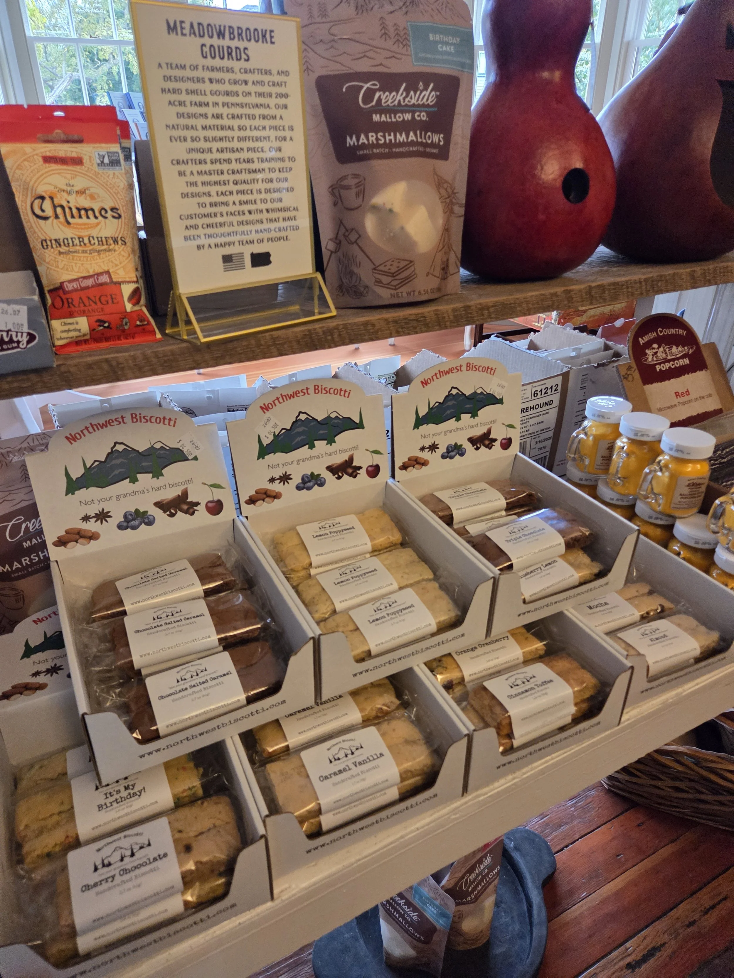 Display shelf with assorted baked goods in boxes labeled Northwest Biscotti, featuring flavors like lemon, caramel vanilla, cherry almond, and mocha, at a small retail shop.