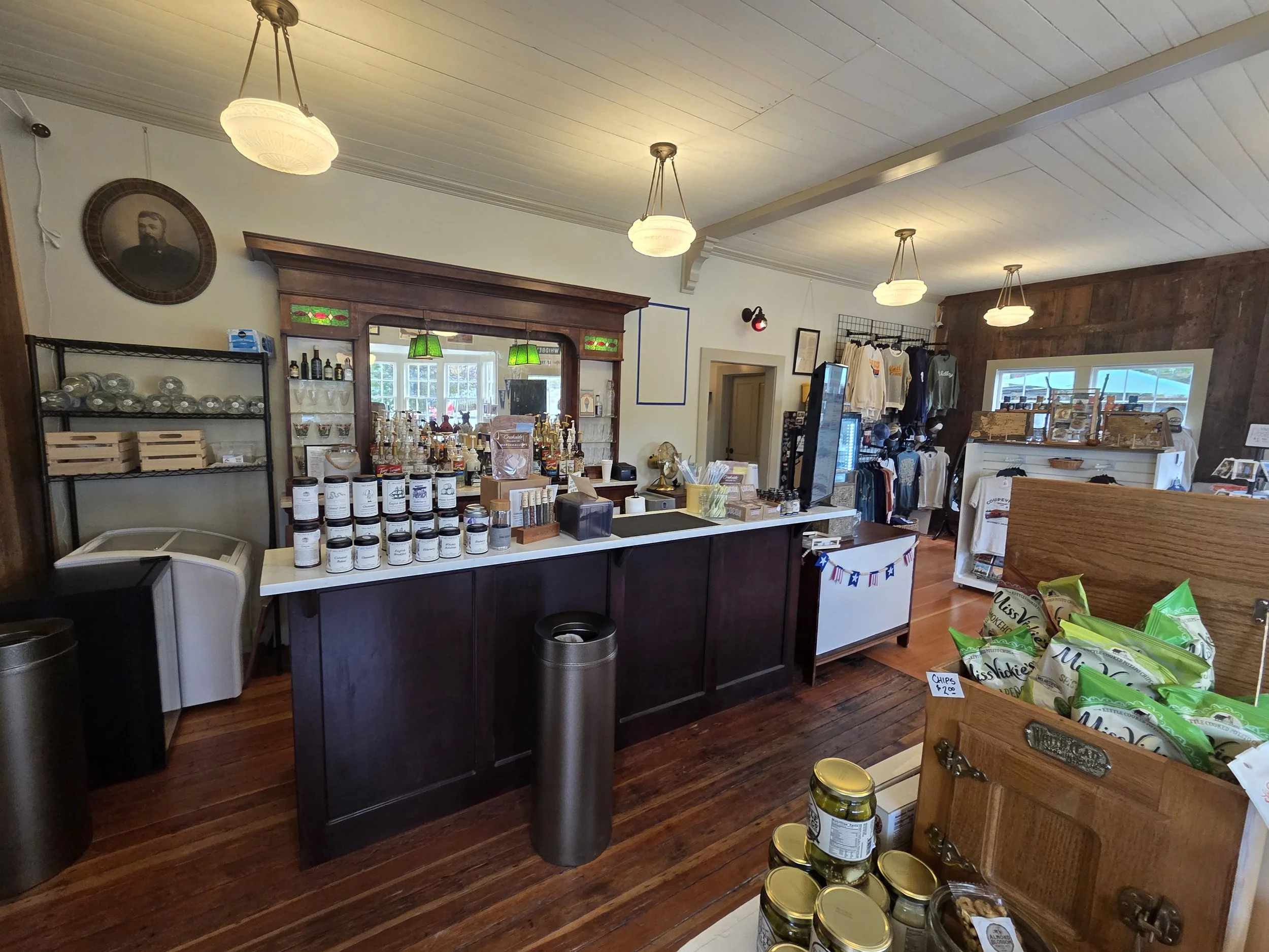 Interior of a small retail store with wooden flooring and ceiling. Features include pendant lights, a display counter with products, a wall with clothing and accessories, and a vintage portrait framed on the wall.