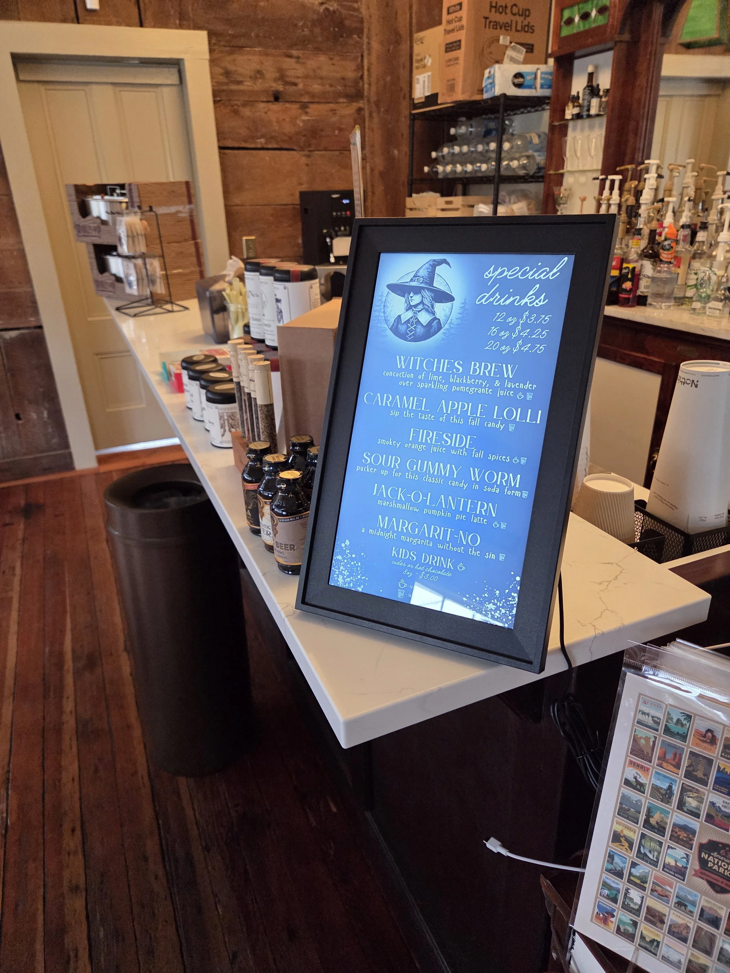A display counter featuring various bottles of syrup or flavoring, with a framed menu sign showcasing the drink options such as Witch's Brew, Caramel Apple Lolli, Fireside, Sour Gummy Worm, Jack-O-Lantern, and Margarita-No, inside a rustic wooden room.