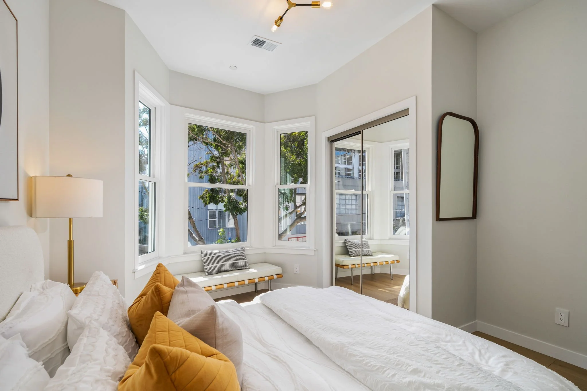 A bedroom with large windows, a bed with white bedding and yellow and gray pillows, a tall mirror near the window, and a modern ceiling light fixture.