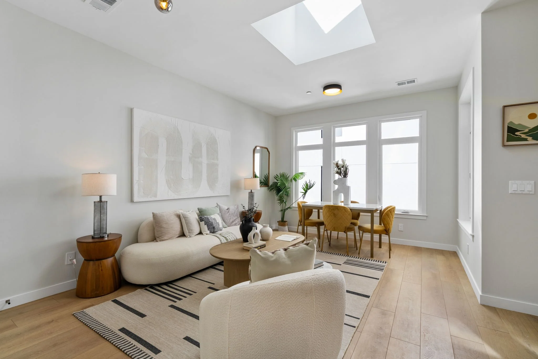 A bright and modern living room with minimalist decor, a beige sofa with plush cushions, a wooden side table with a lamp, a coffee table with decorative vases and books, a patterned rug, and a dining area with a white table and yellow upholstered cha
