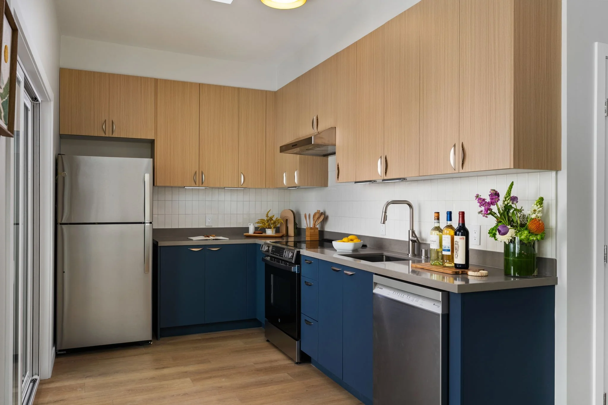 Modern kitchen with wooden upper cabinets, blue lower cabinets, stainless steel appliances, a white tile backsplash, and a window with a sliding door. Countertop items include a bowl of lemons, bottles of wine, and a vase of flowers.
