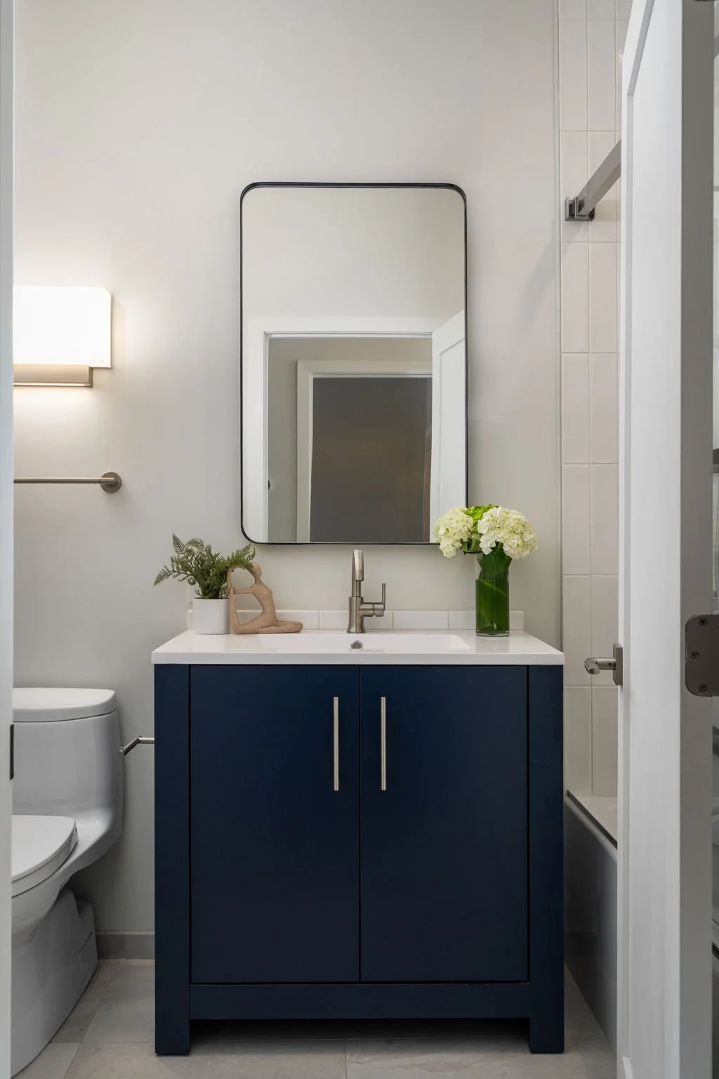 Modern bathroom with navy blue vanity, large mirror, white sink, beige walls, small potted plant, vase with white flowers, and a door leading to a shower.