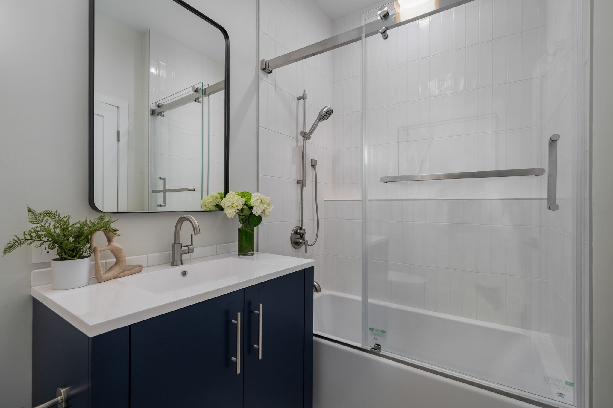 Modern bathroom with a white sink vanity, navy blue cabinets, a large mirror, potted plants on the counter, white tiled shower with grab bars, and a showerhead.