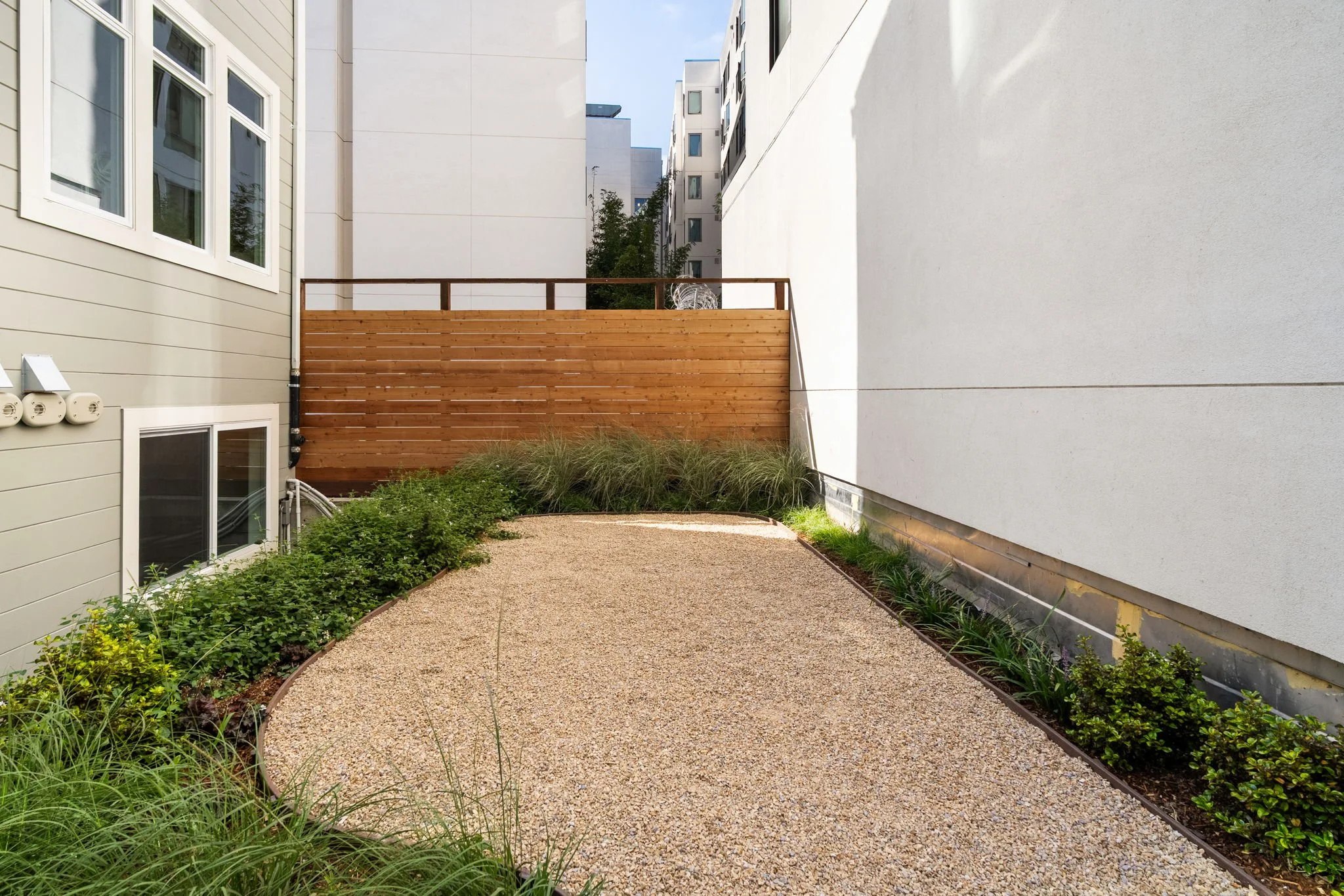 A small urban backyard with gravel pathway, surrounded by plants and bushes, with modern multi-story buildings and a wooden privacy fence in the background.