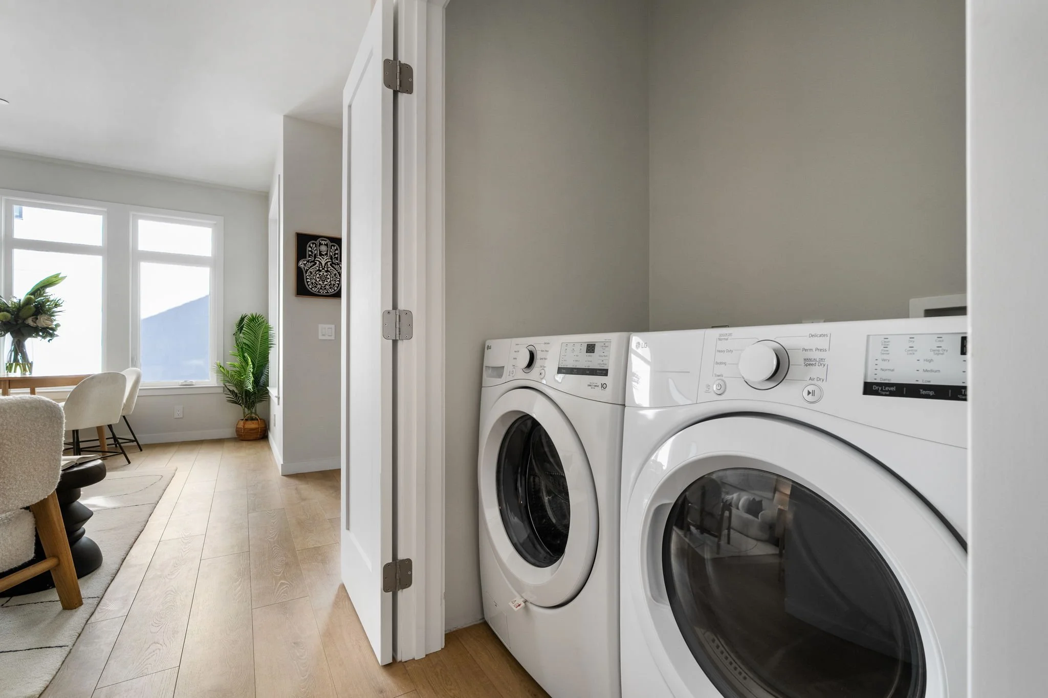 Laundry room with a pair of white LG front-loading washer and dryer, a small wooden dining table with chairs, a window with sunlight, a vase with flowers, and a potted plant.