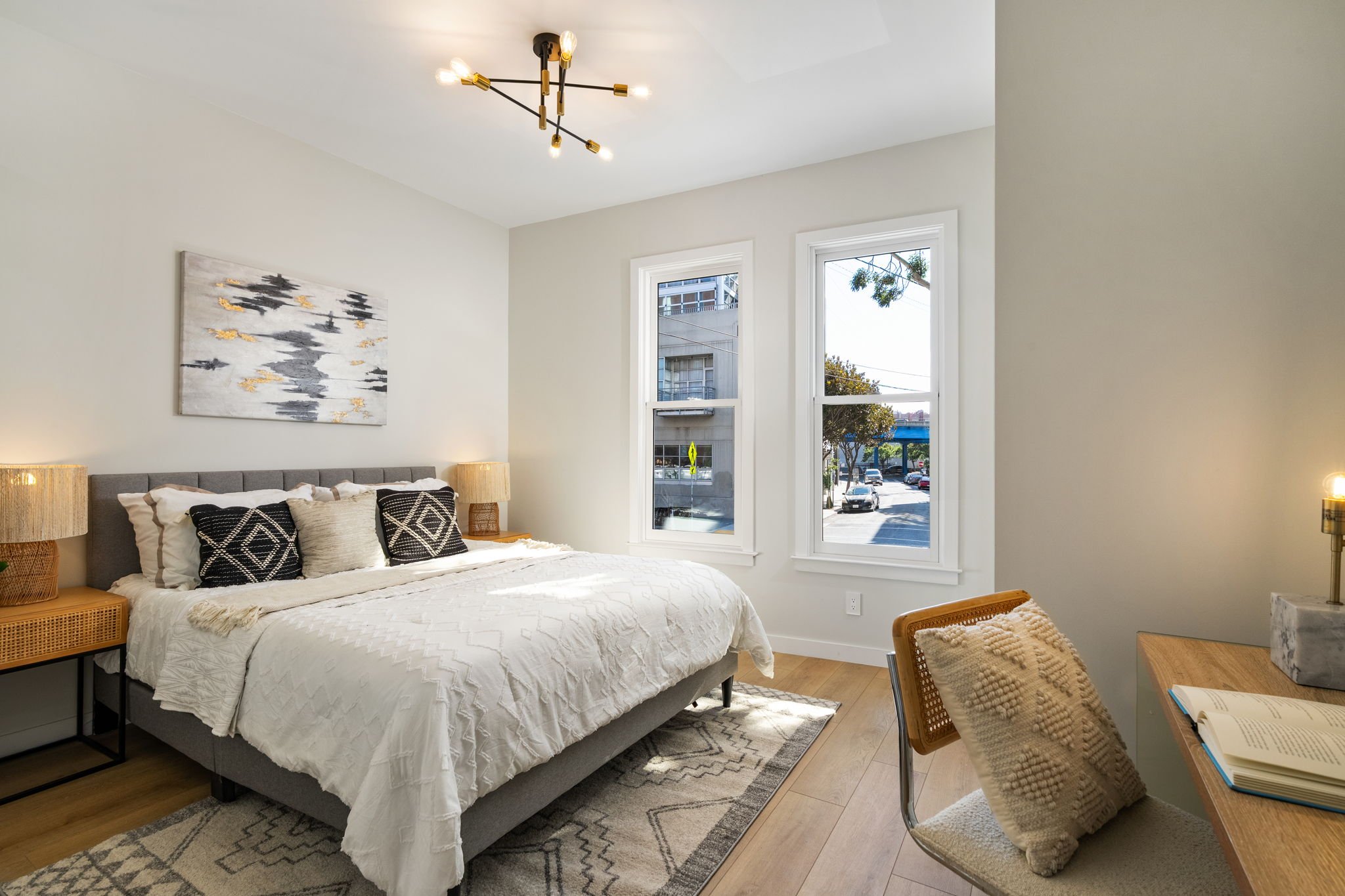 A bedroom with a gray upholstered bed, white bedding, and black-and-white decorative pillows. Two wooden nightstands with woven lamps are on each side of the bed. A large abstract painting with black, gray, and gold accents hangs above the bed. Two l