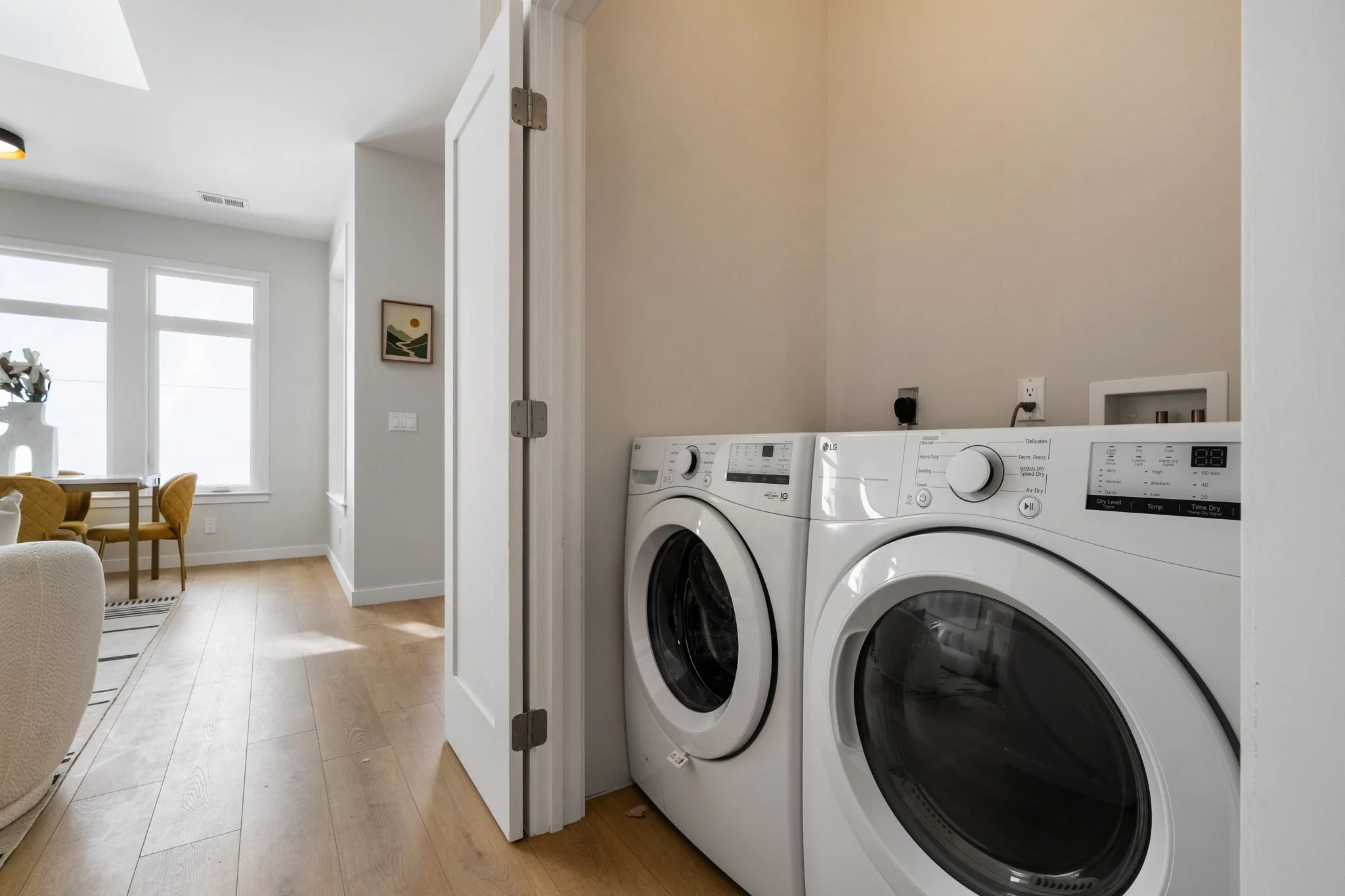A laundry area in a house with a white washer and dryer side by side in a small space, with a wall outlet and electrical switch nearby, adjacent to a living and dining area with large windows, a table, chairs, and decor in the background.