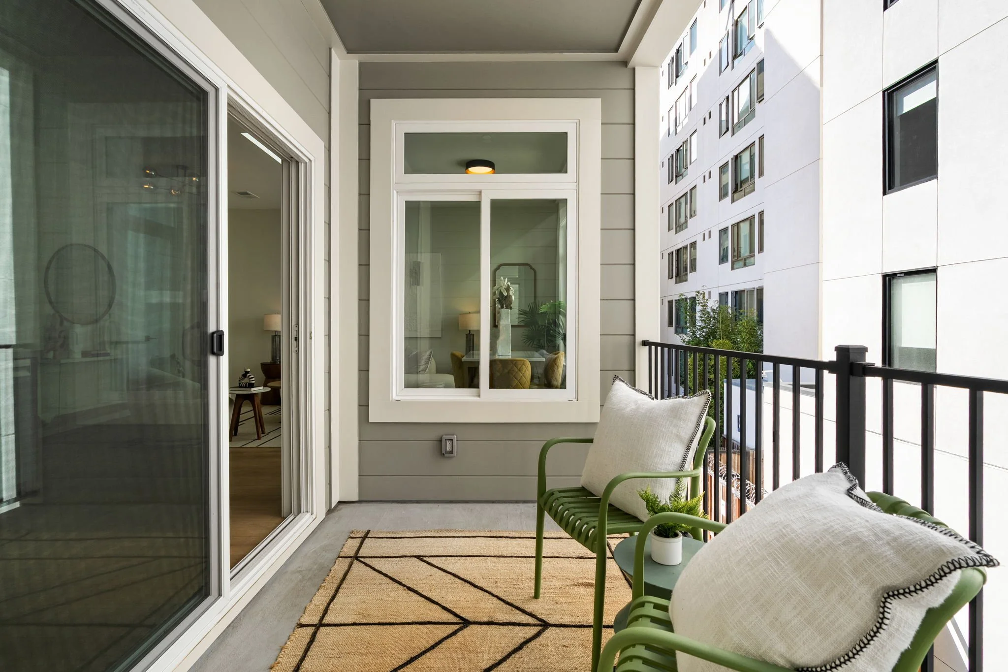 Balcony with green metal chair, beige cushion, small potted plant, striped rug, sliding glass door, and city apartment building in the background.