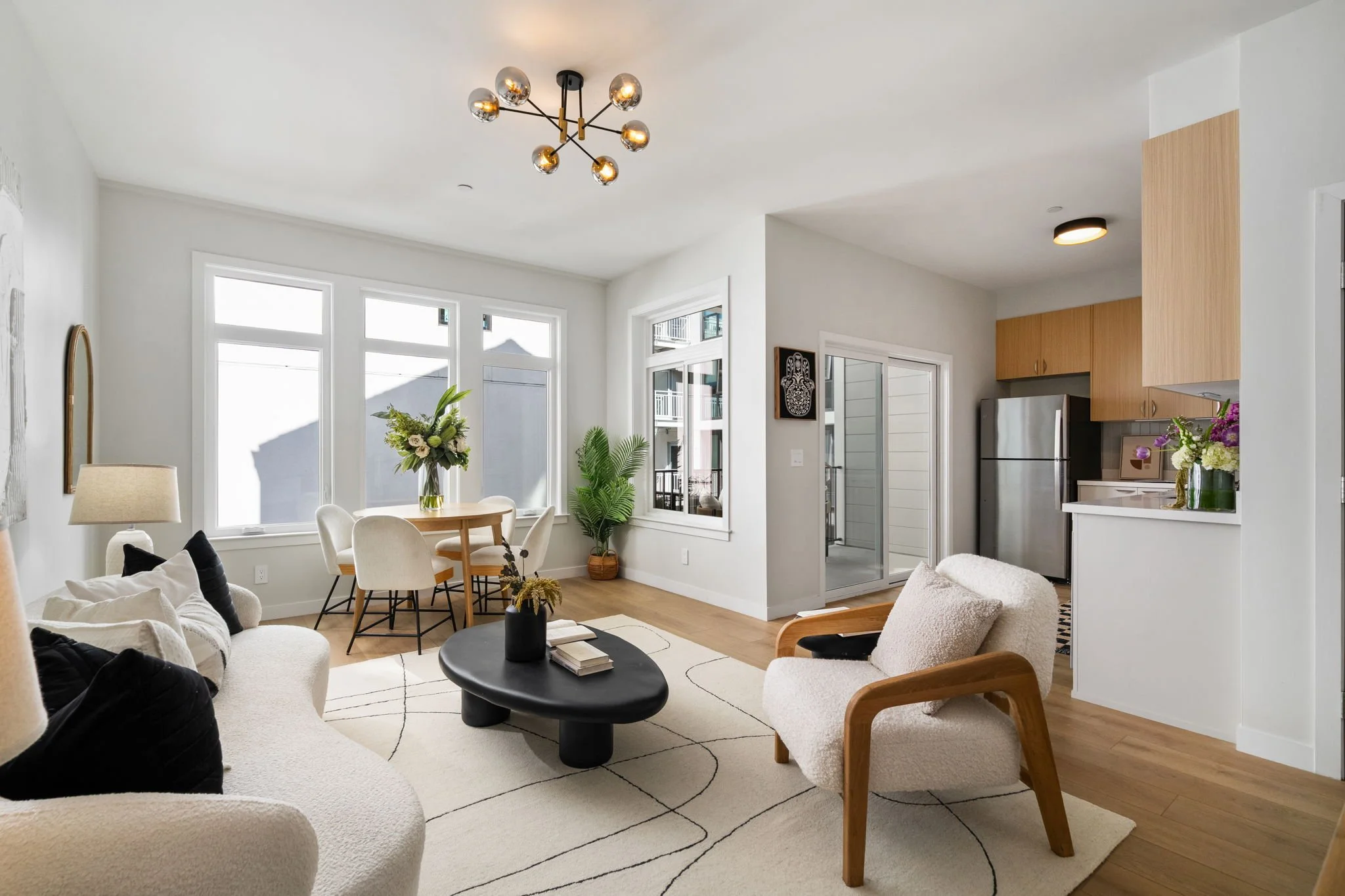 Modern living room with white walls, large windows, wooden flooring, a black oval coffee table, white and black sofas, beige armchairs, a round dining table with chairs, green plants, fresh flowers, and contemporary light fixtures.