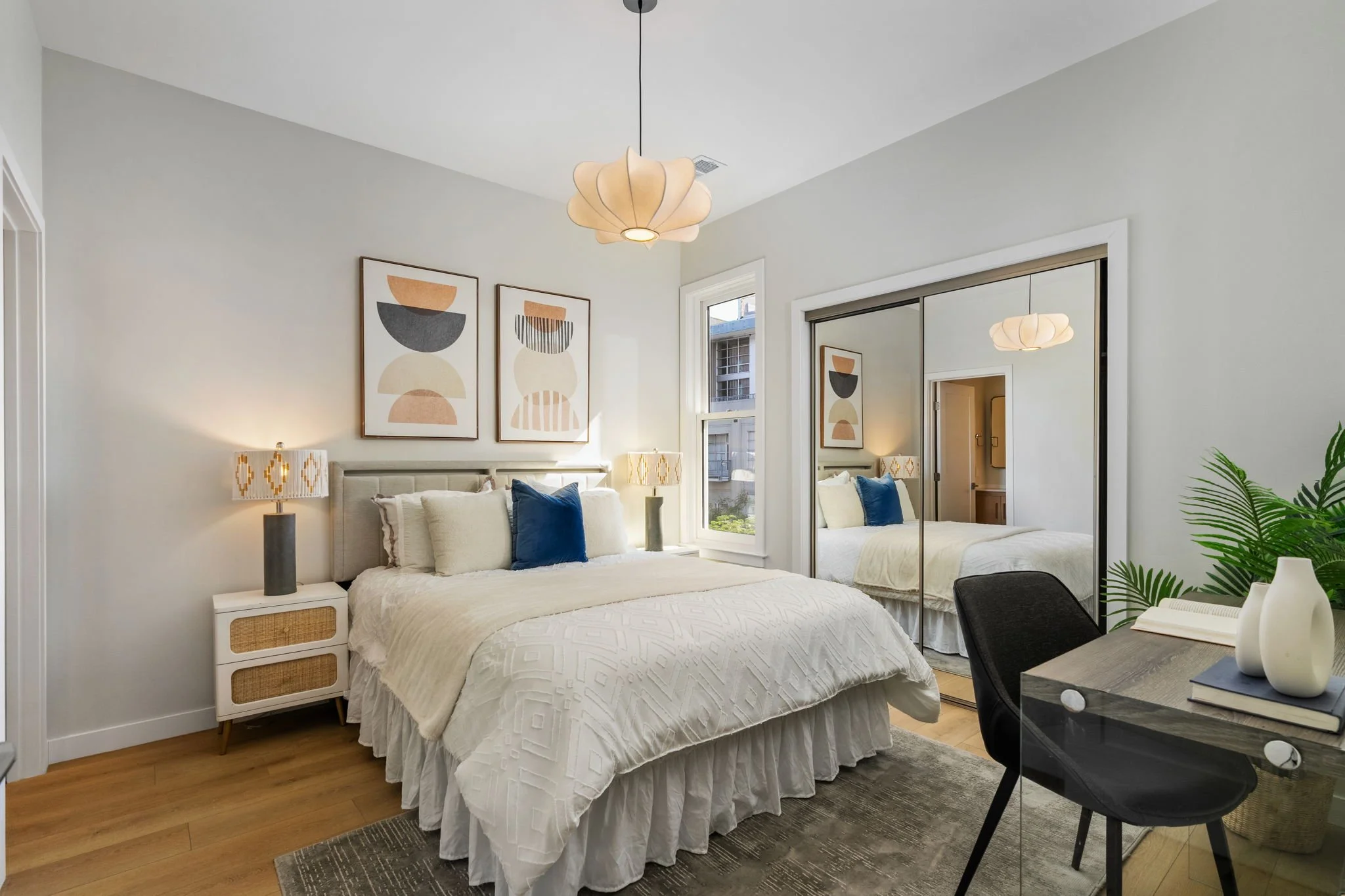 Modern bedroom with white bedding, blue pillows, wooden flooring, abstract art, round bedside lamps, a large mirror closet, and a chair next to a glass table with decorative vases.