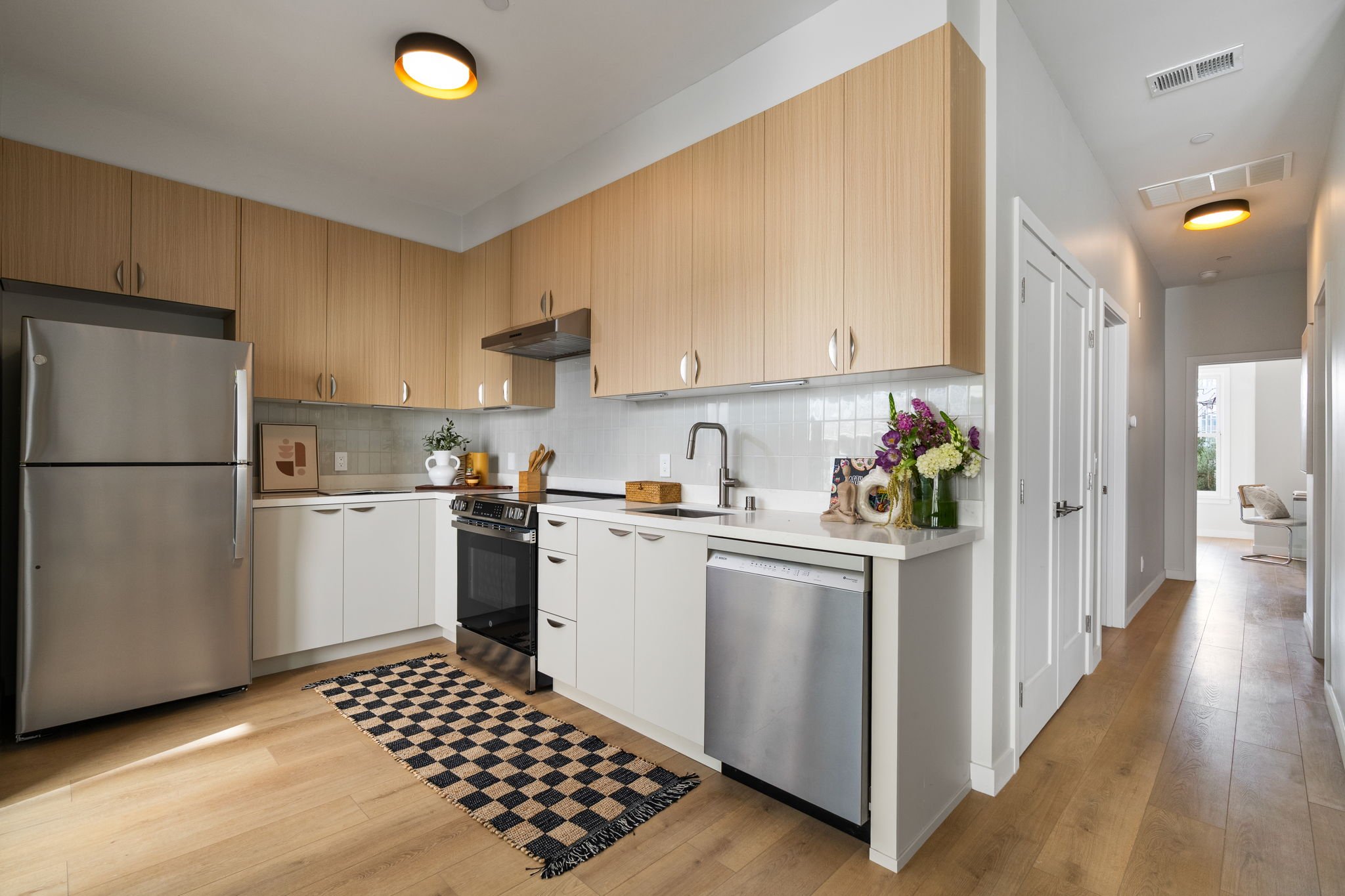 Modern kitchen with white and wood cabinets, stainless steel appliances, a checkered rug, and a vase of flowers on the counter near the sink.