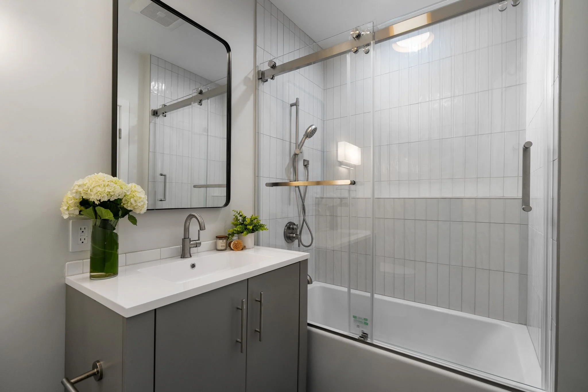 Modern bathroom with white tiled shower area, glass door, and a dark vanity with a white countertop. There is a large mirror above the sink, a vase of white hydrangeas, and some toiletries on the countertop.