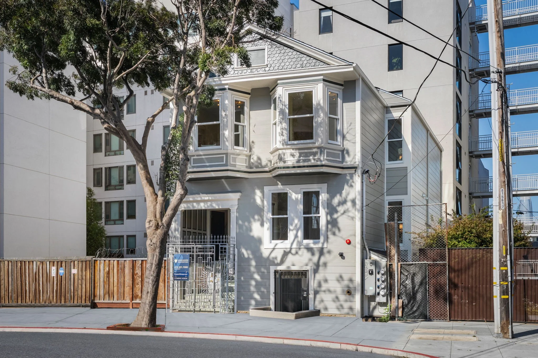 A three-story residential building with a white facade, bay windows, and detailed moldings on the front exterior. A large tree partially obscures the building, and there is a wooden fence at the sidewalk. Utility poles and wires are visible on the right side of the image.