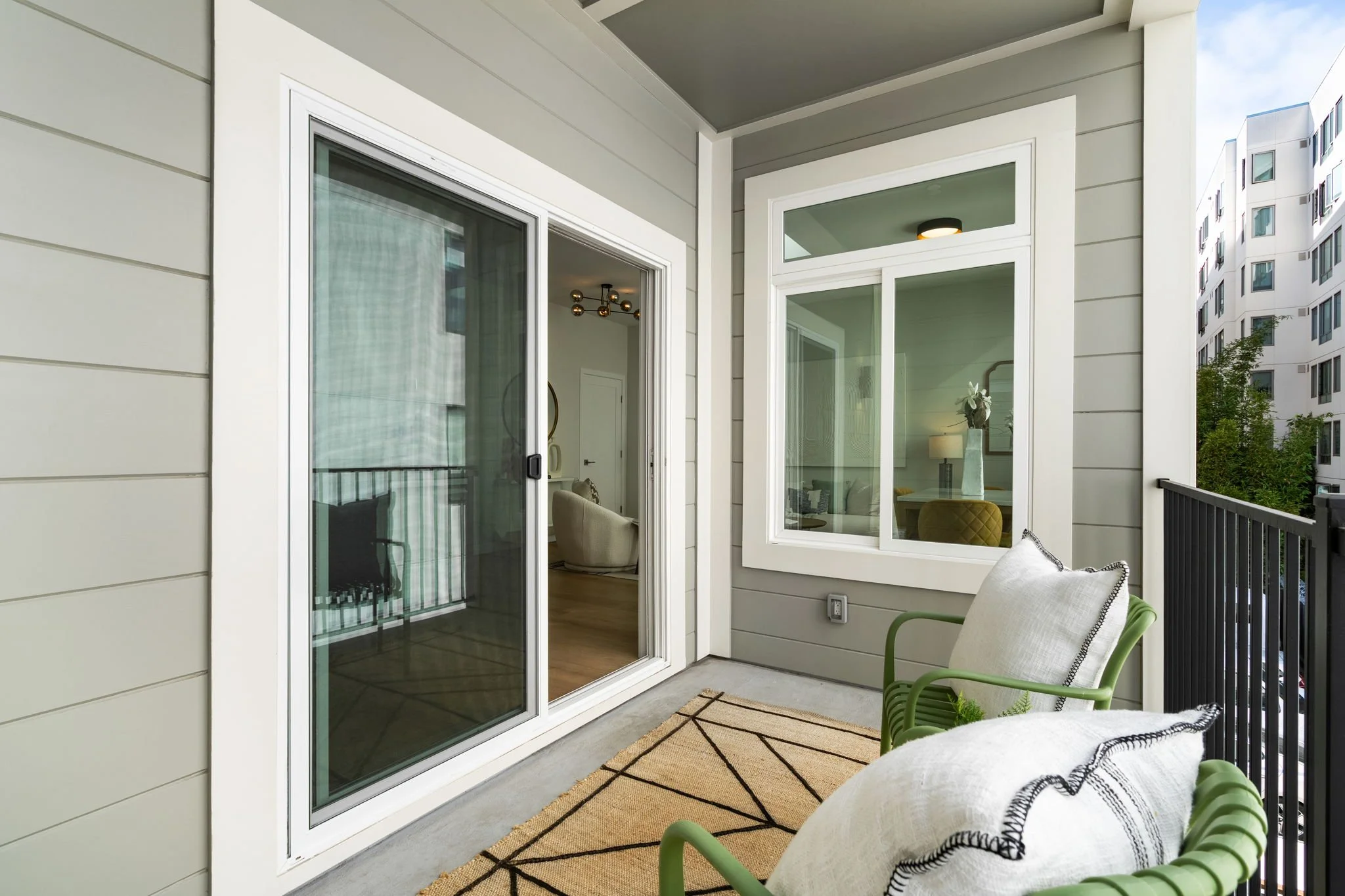 Balcony with a beige chair and a pillow, a beige rug with black geometric patterns, and sliding glass doors leading into a modern apartment with visible interior furniture.